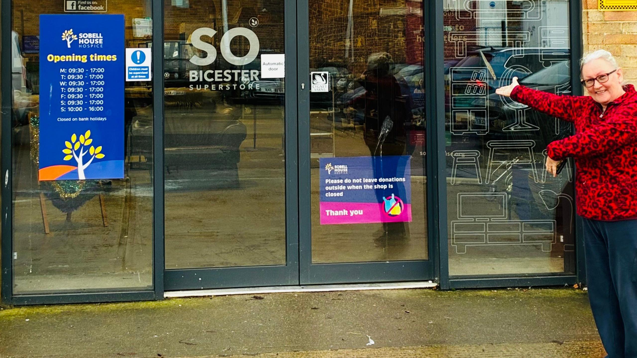 A smiling Jay Boydon outside Sobell House shop in Bicester. She is looking at the camera and is pointing towards the entrance. The ground in front is wet.