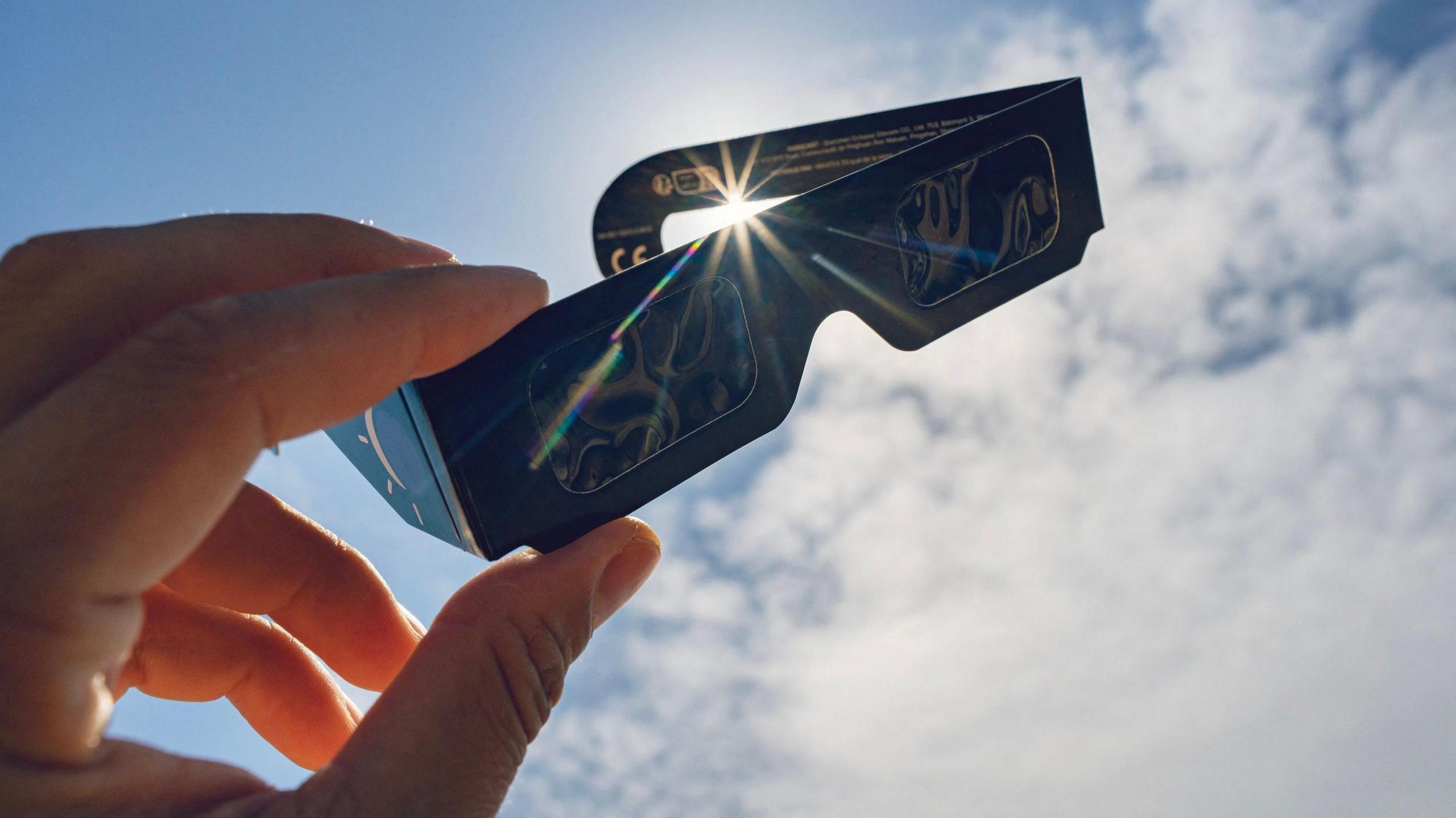 Hand holding up eclipse-viewing glasses to a blue sky with a few white fluffy clouds