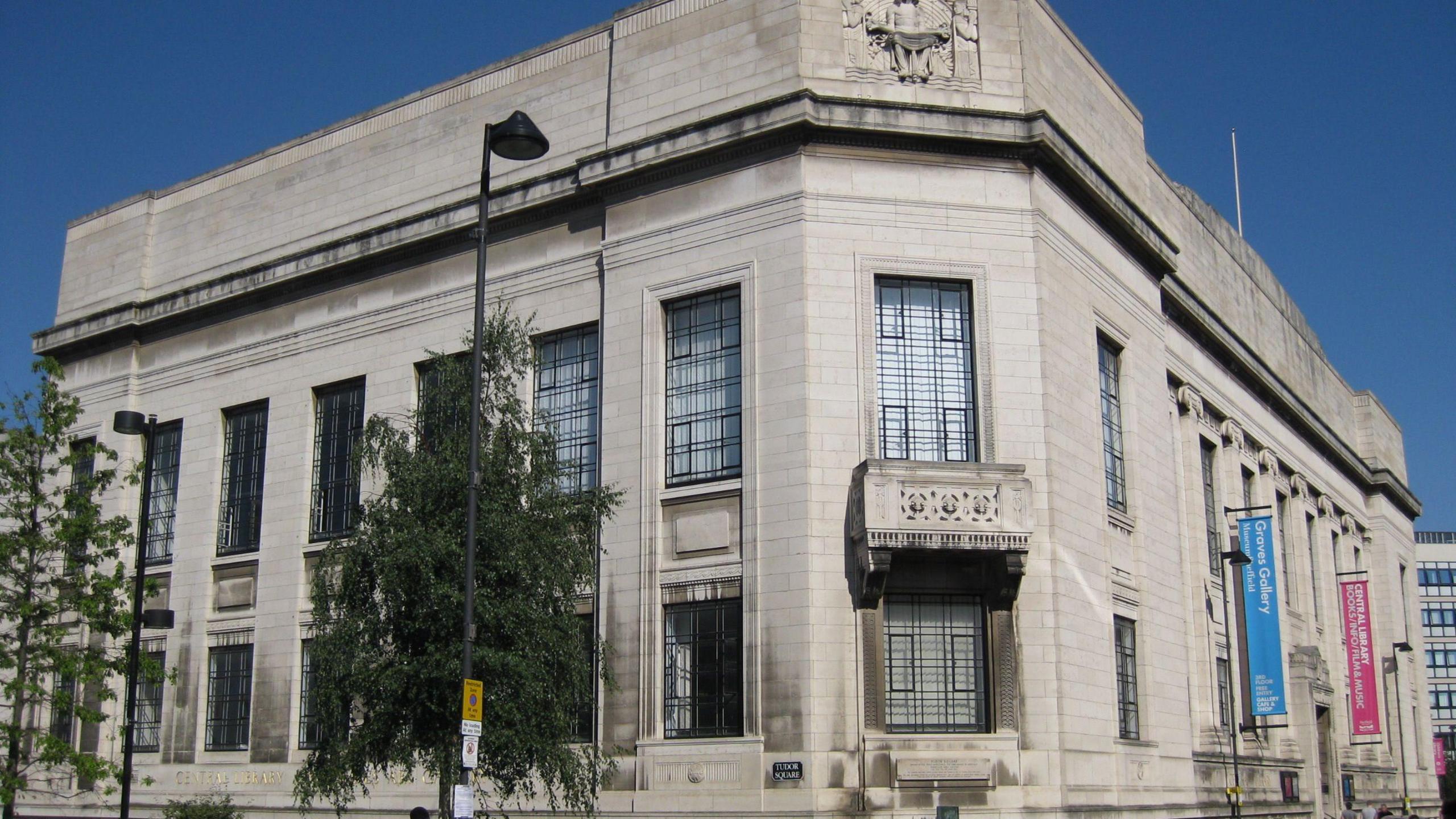 A large white brick building covers a corner of a street. There is a balcony, large windows and colourful banners hanging from one wall