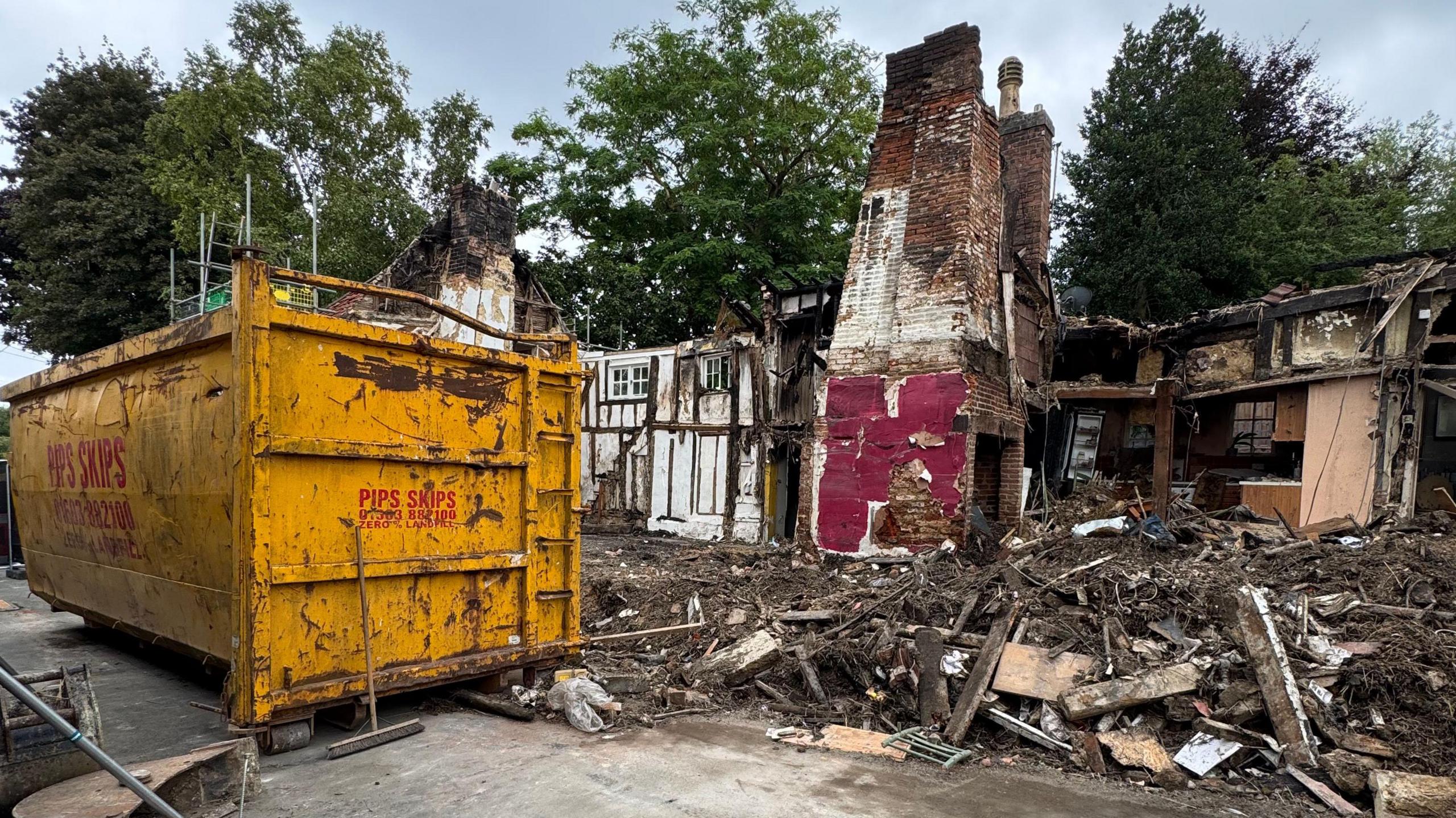 A large yellow skip next to a row of destroyed cottages and large piles of rubble and debris