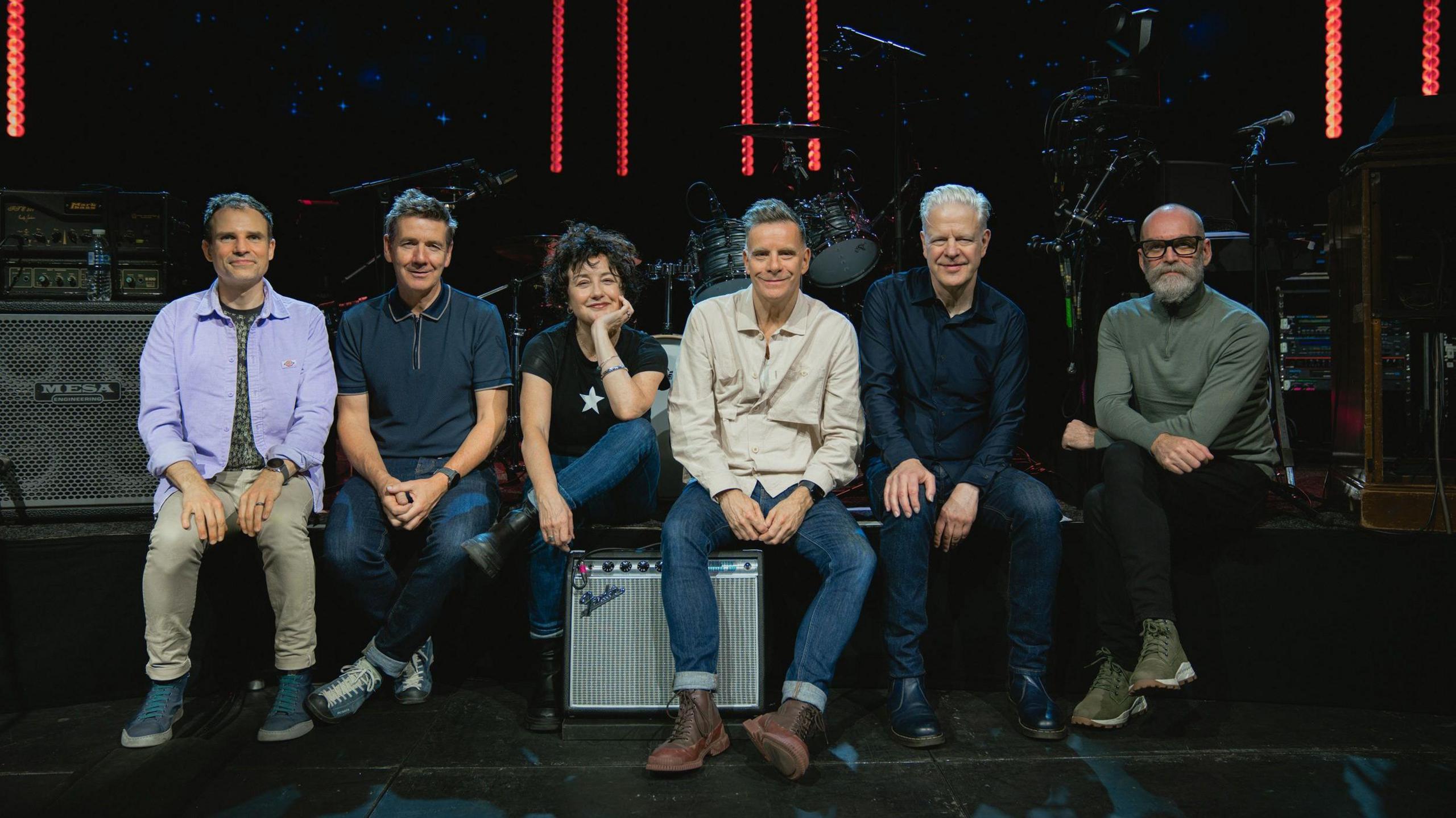 A group of six people sat on a dark stage and looking at the camera. There are five men and one woman.