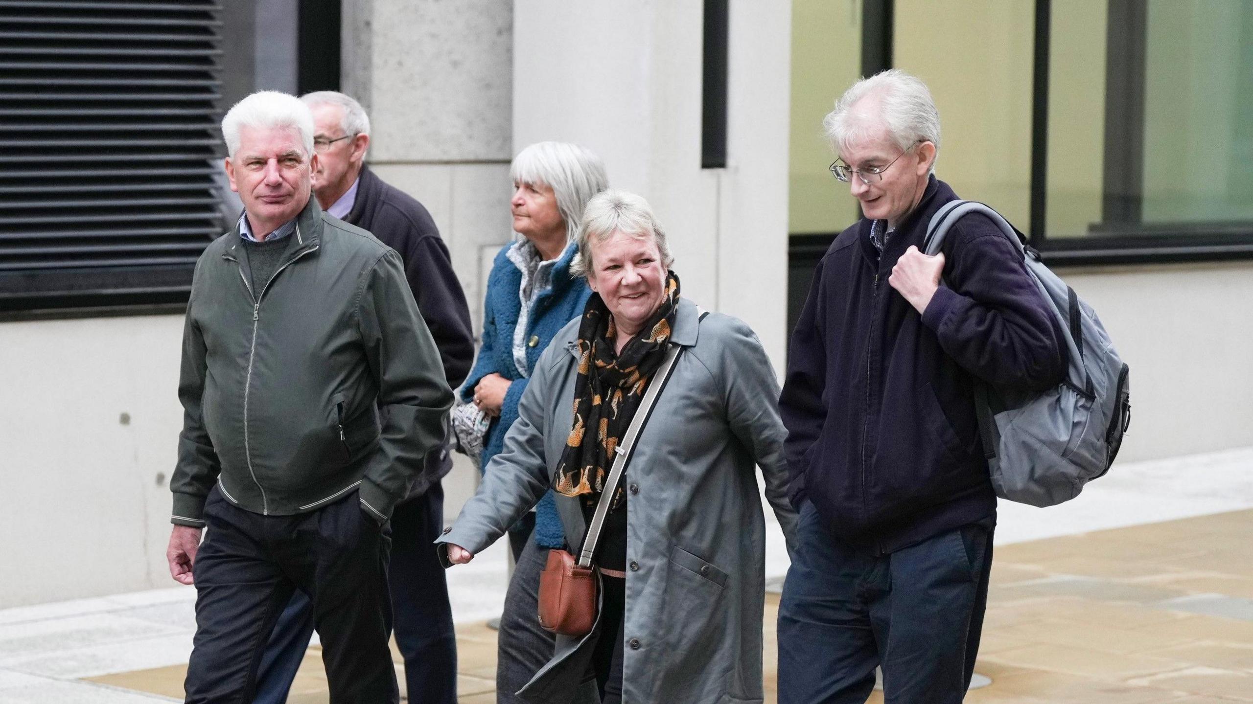 A group of three men and two women, all with grey hair, walk together outside a building. 