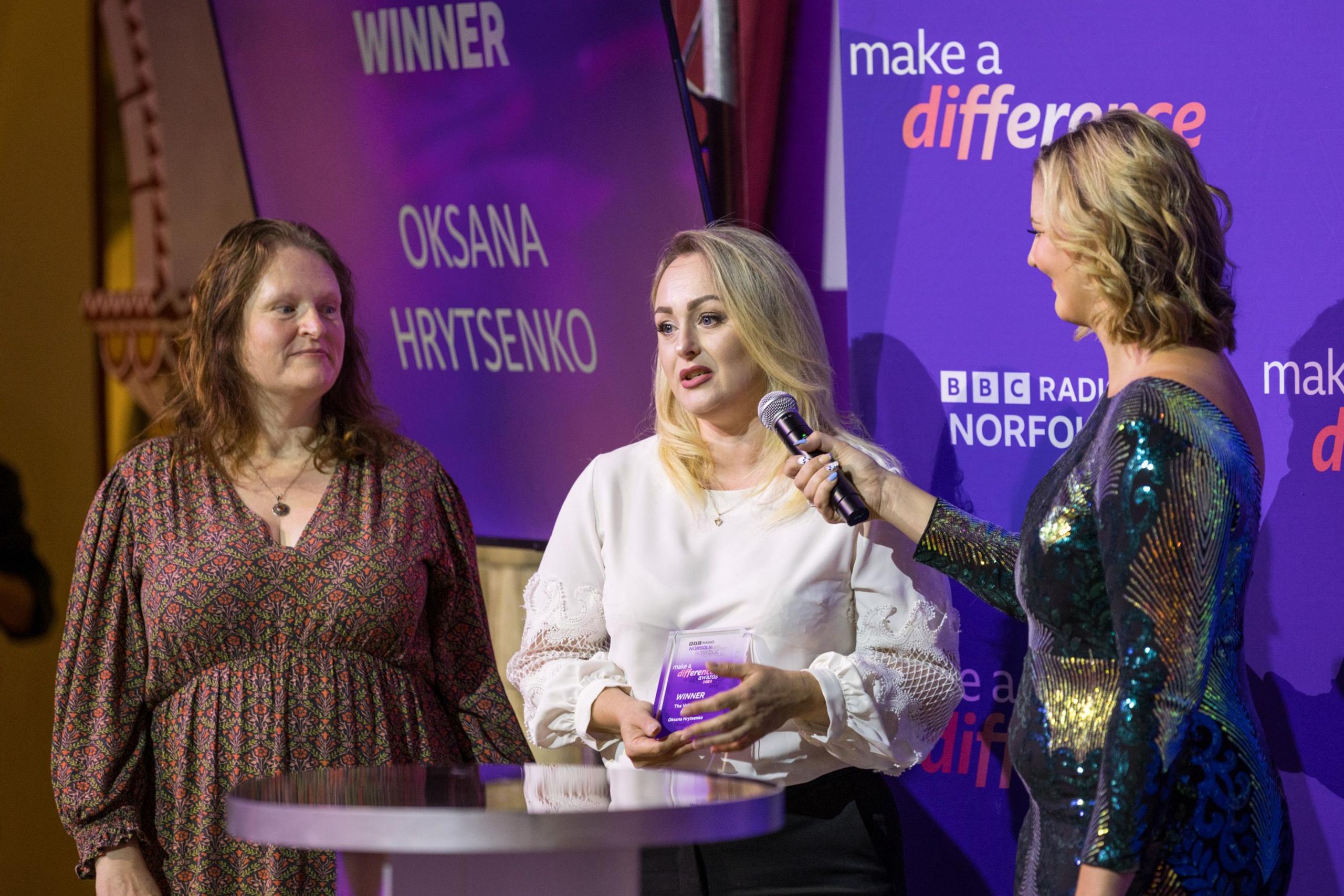 Three women stand on a stage in front of a purple background. The woman in the middle has blonde hair, is wearing a white shirt and is speaking into a microphone. She is holding a glass award. 
