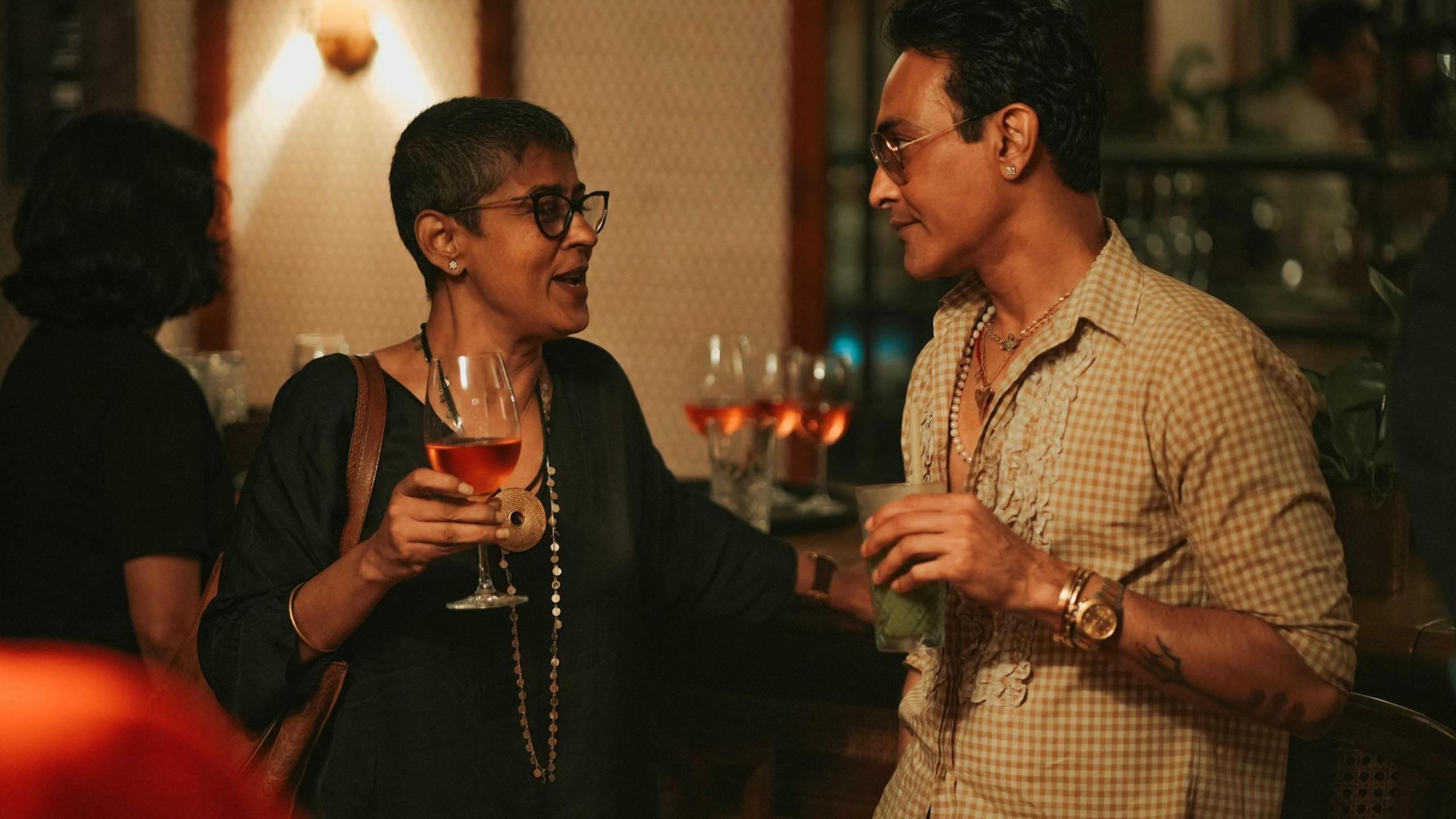 A man and a woman at a club having a drink and engaged in an animated chat. The man is wearing a checked shirt with sunglasses while the woman is wearing a black dress with glasses. 