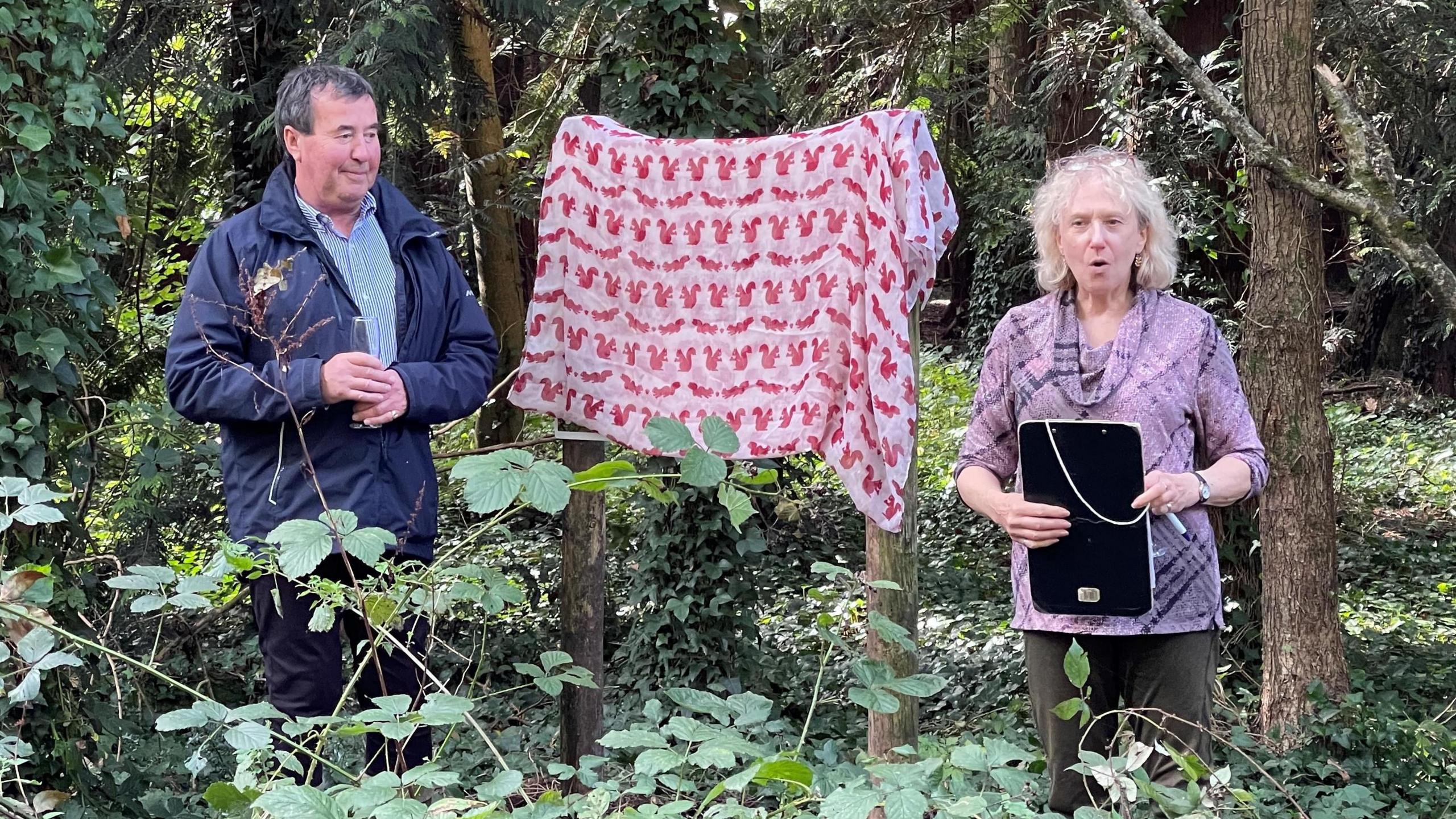 Simon Howes, a man wearing a navy blue coat, and Helen Butler, a woman with short blonde hair, stand next to a sign in woodland. The sign is covered in a red blanket with squirrels on