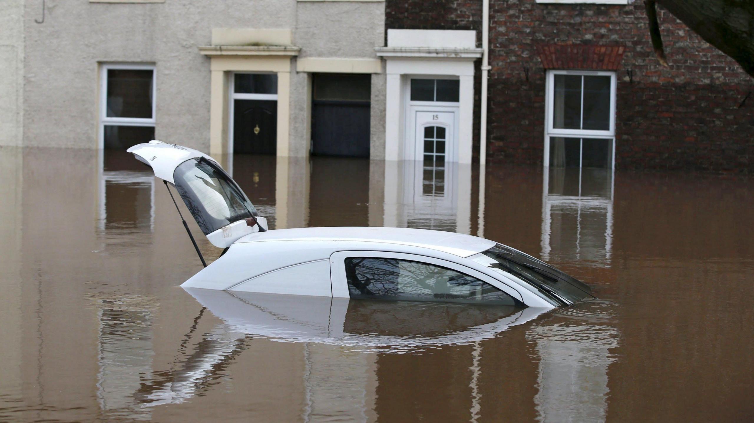 A flooded street in Carlisle, Cumbria, following Storm Desmond in 2015. There is a white car, mostly submerged, with its boot open, in the foreground, with terraced houses behind. The upstairs windows have yellow crosses marked on them.