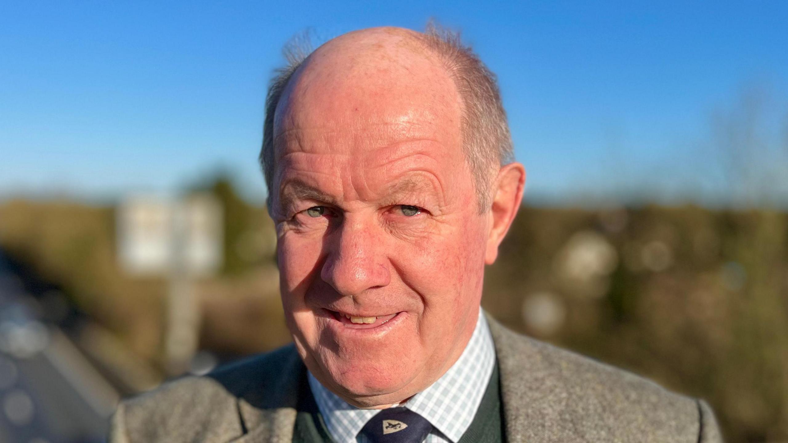 Tim Passmore smiles at the camera as he stands on a bridge overlooking a dual carriageway on a sunny day. He wears a tweed grey suit jacket, white and blue chequered shirt, green jumper and a tie. He has short grey hair which has thinned on the top of his head. 