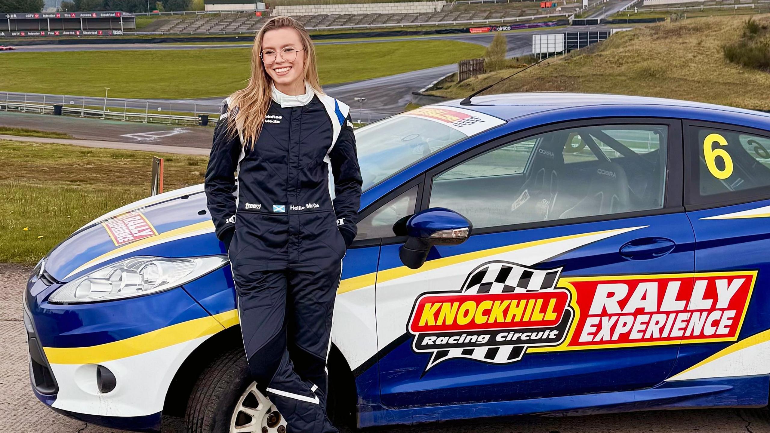 Hollie McRae in a racing suit standing next to a blue and white rally car. She has blonde hair and is wearing glasses, smiling at the camera. The car has a "Knockhill Racing Circuit" logo and the number 6 on the side. The background shows a racetrack and grassy areas.