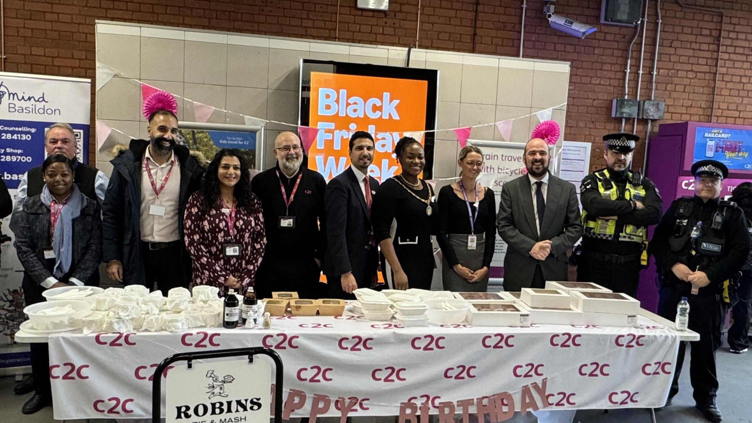 Dignitaries and railway staff standing in front of a table which has food on it. They are inside the station building. There are 11 people posing for the photo, including c2c staff, Conservative MP Richard Holden and two police officers