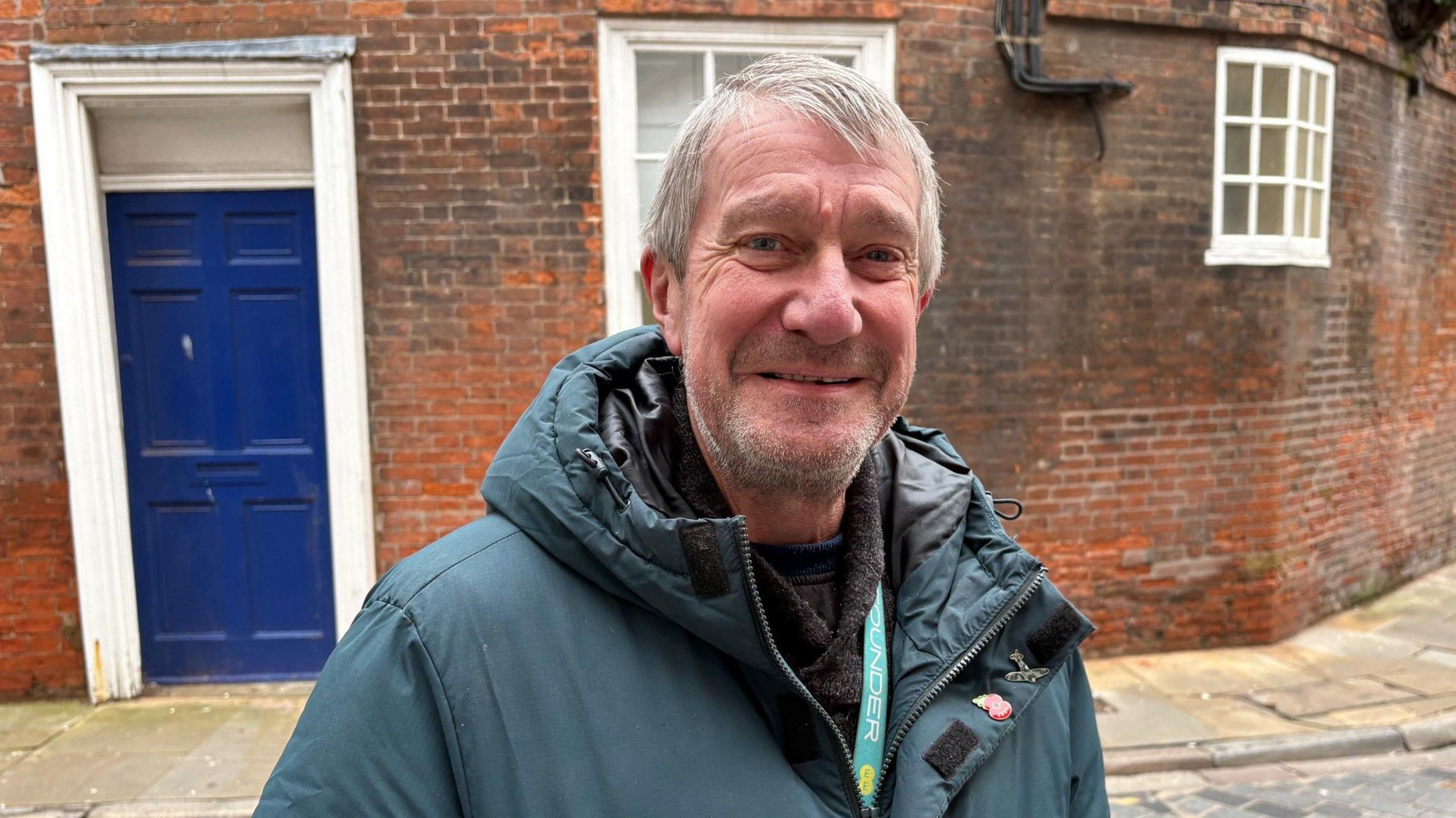A man in his 60s, with grey stubbles, is posing for the camera in front of a red brick corner home with a blue door.