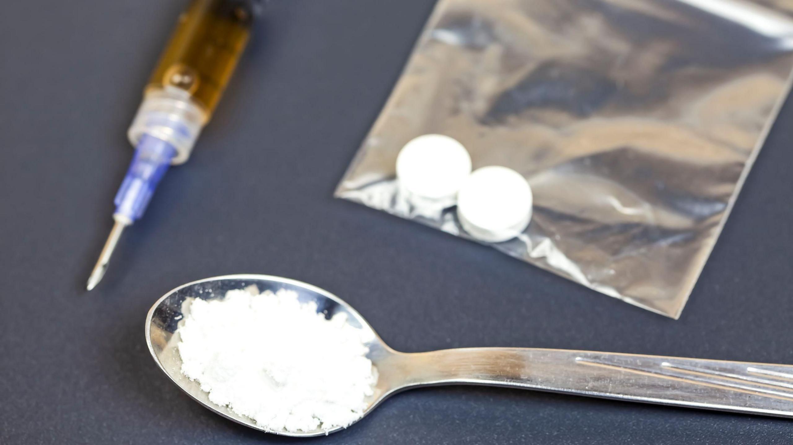 On a table are various items including a syringe, a spoon with powder on it and a clear plastic bag with two pills inside.