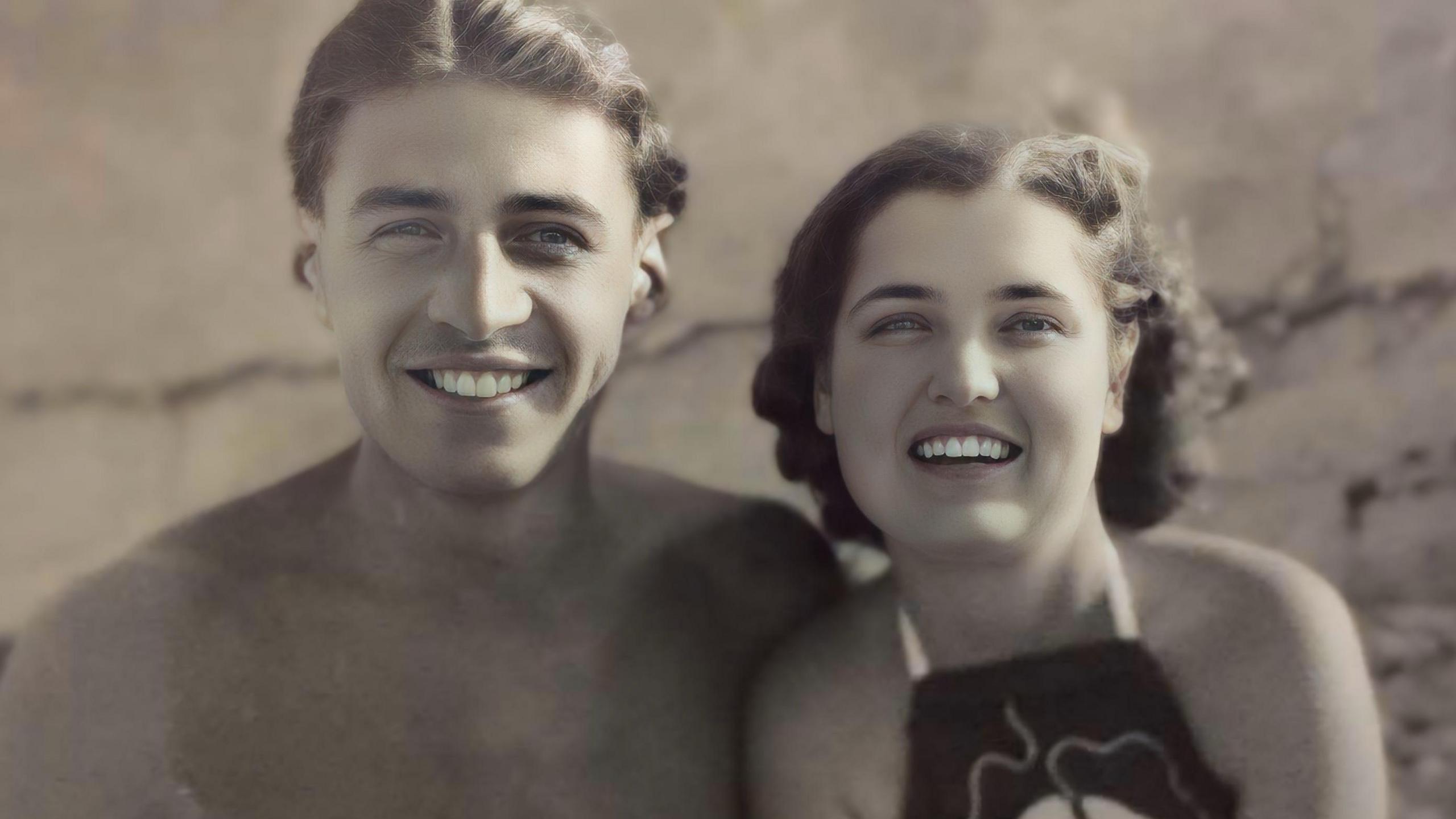 A black and white image of a young couple smiling at the camera. The man with short dark hair with a centre parting is on the left of the picture, topless on the beach. The young woman beside him has shoulder length curly dark hair and is wearing a dark coloured halter-neck swimming costume.