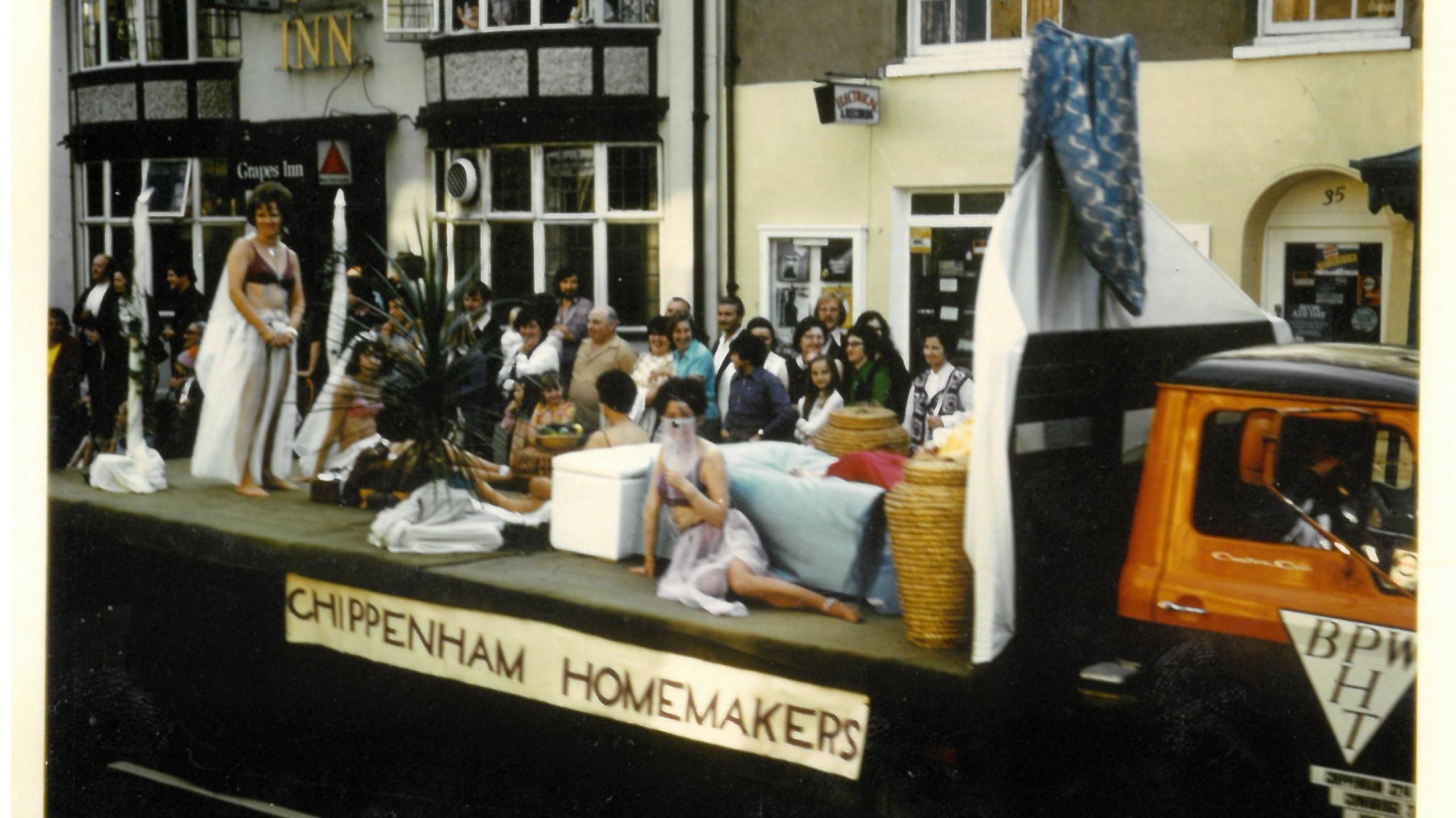 A group of women lie and stand on the back of a lorry with a sign saying 'CHIPPENHAM HOMEMAKERS' on the side of the vehicle.