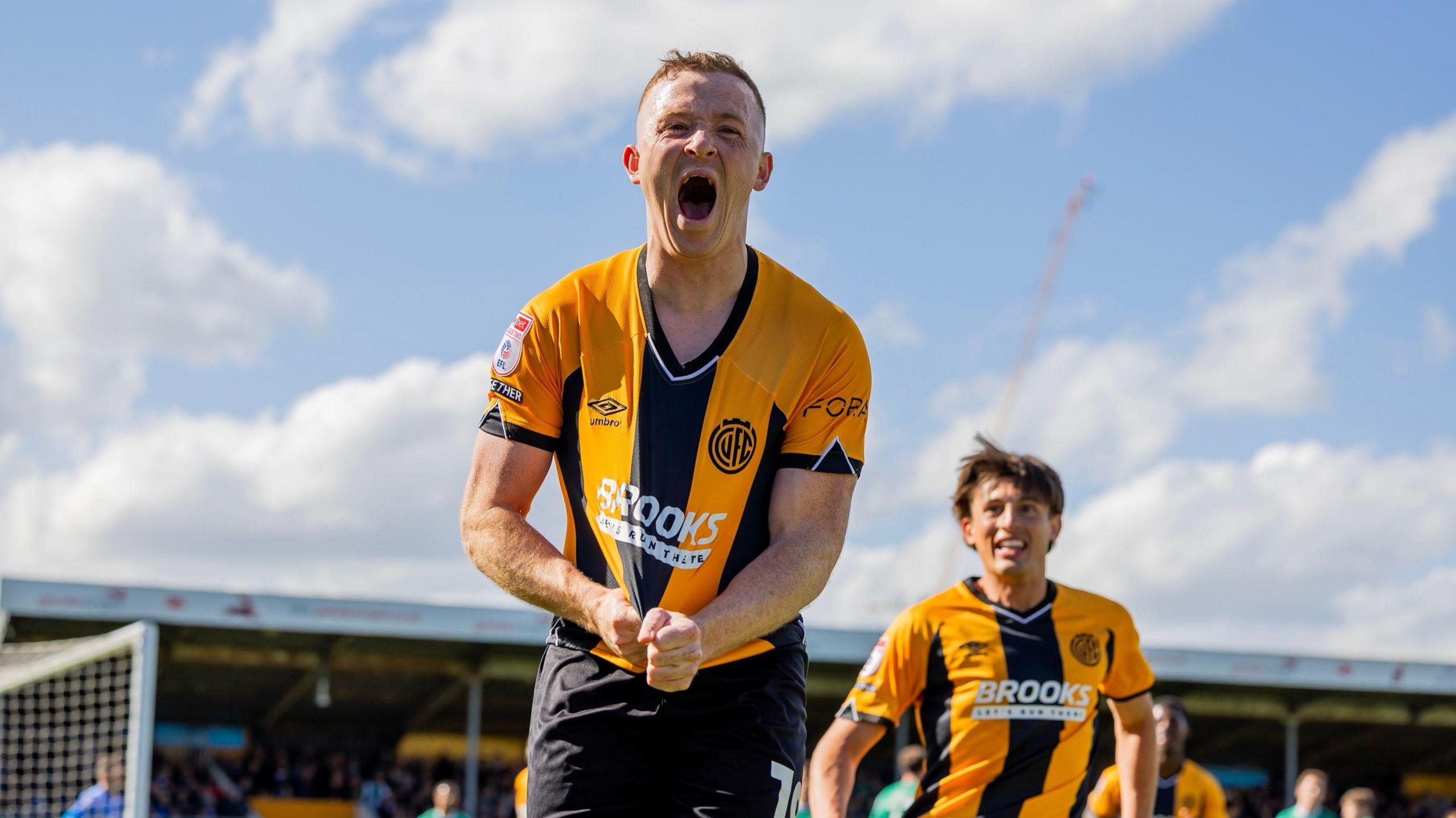 Shayne Lavery pulling at his oragne and black striped Cambridge United shirt as he celebrates his goal against Notts County in May