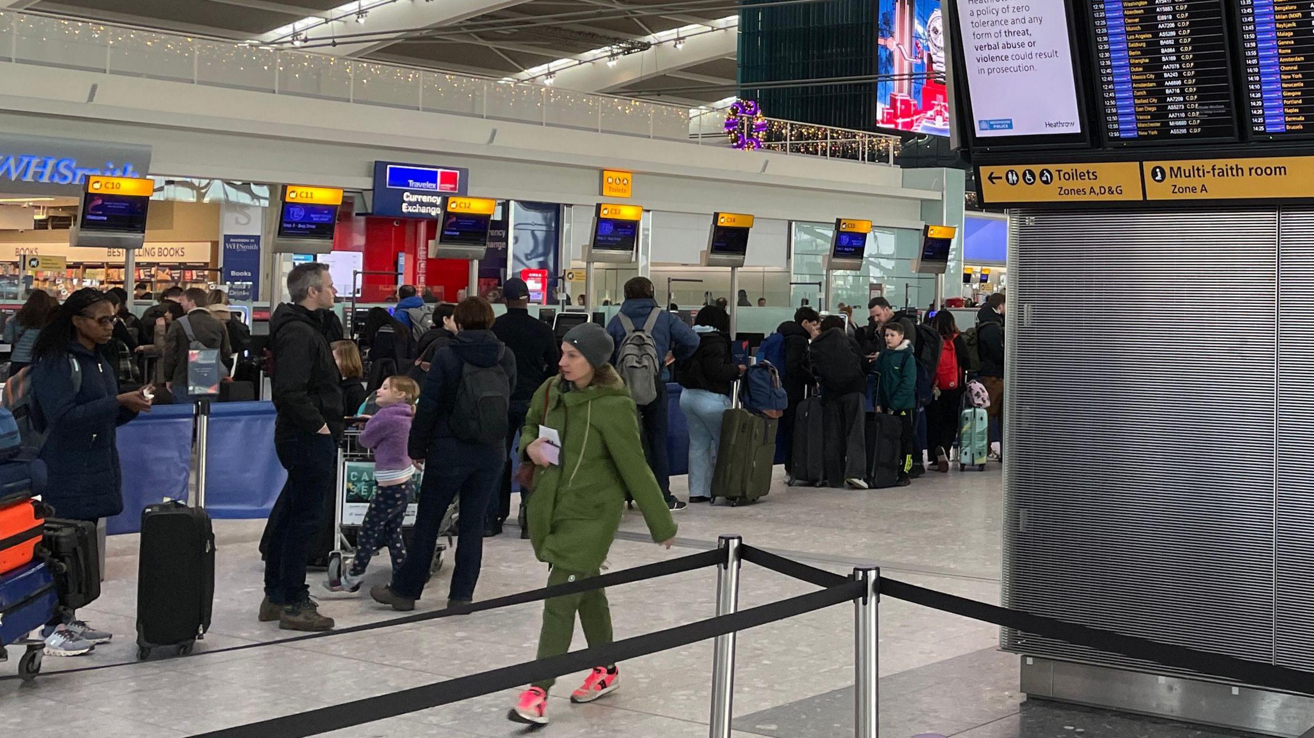 People in coats and with suitcases stand in a line in a Heathrow Airport terminal. A woman in a green coat and green trousers walks in the opposite direction