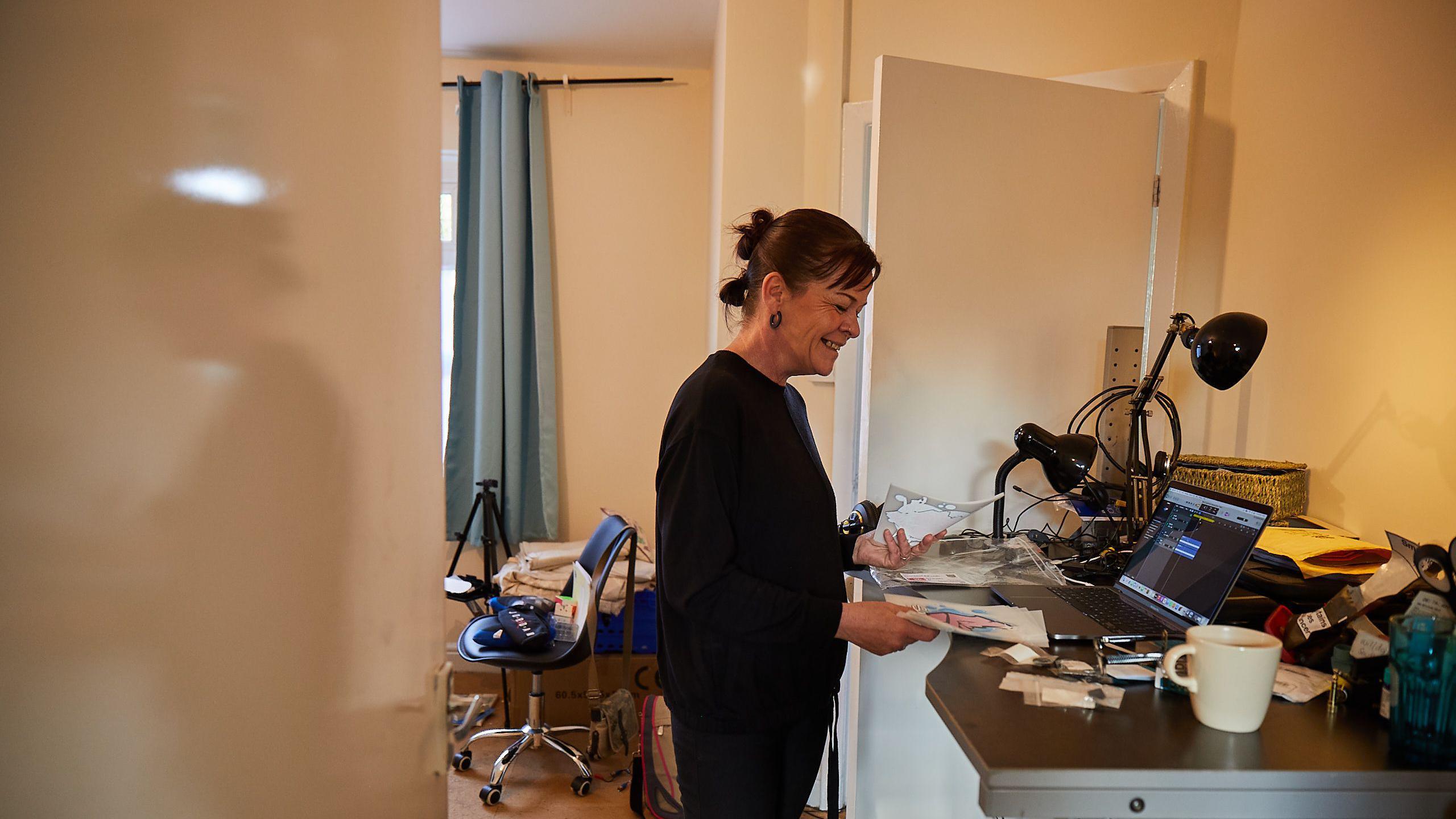 Amanda Wall standing at a desk in her house with a laptop and paperwork. She is wearing a black top and trousers. She has dark hair and black hoop earrings. She is holding paperwork, looking down at it and smiling. 