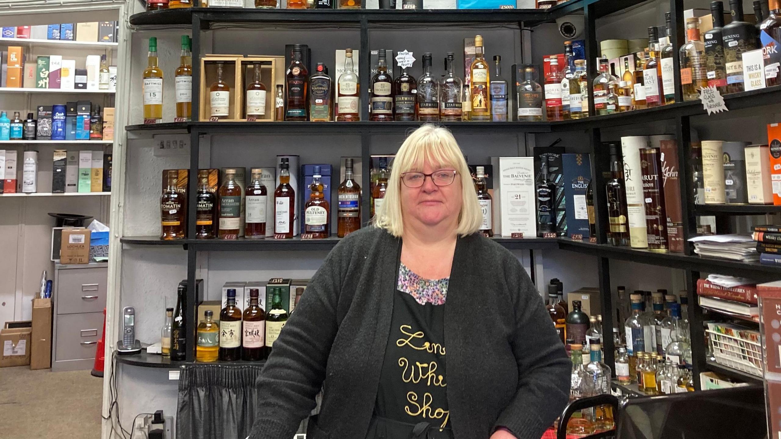 A blonde-haired Shelley Williams standing in front of a row of shelves filled with whisky