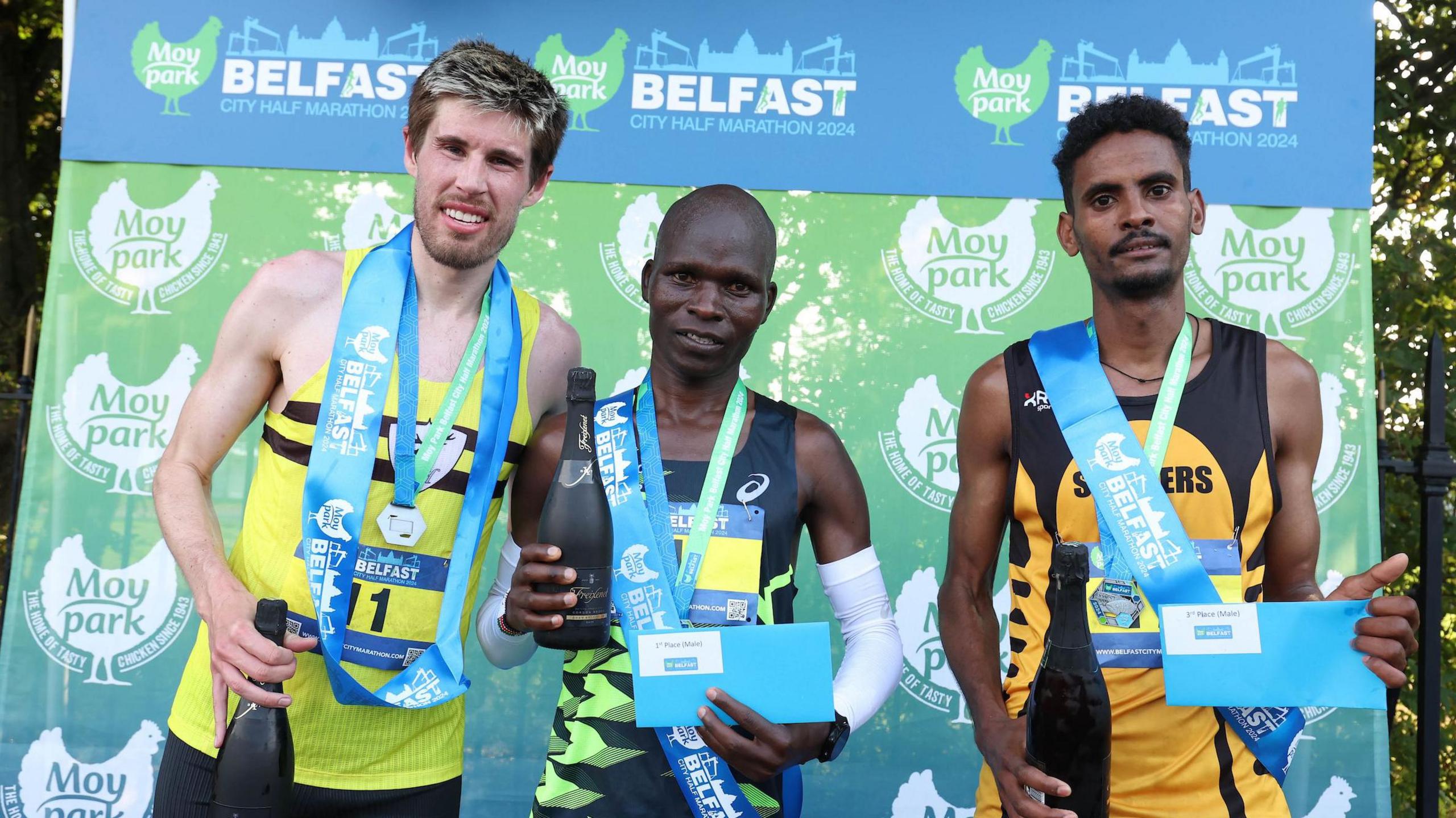 Three men standing on the podium with bottles of champagne. They are each wearing their medals