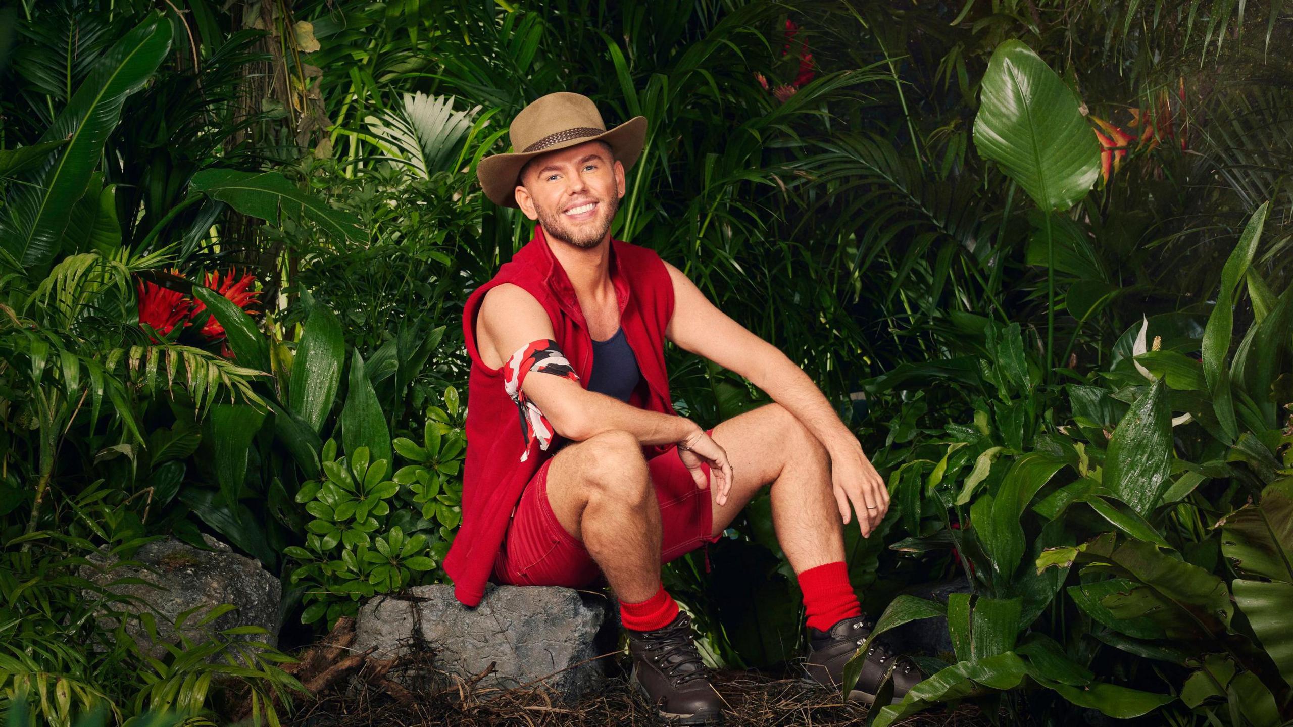 A radio one DJ sitting on top of rocks dressed in a red body warmer, red shorts and red socks. He is wearing a brown hat and is pictured beside a jungle. 