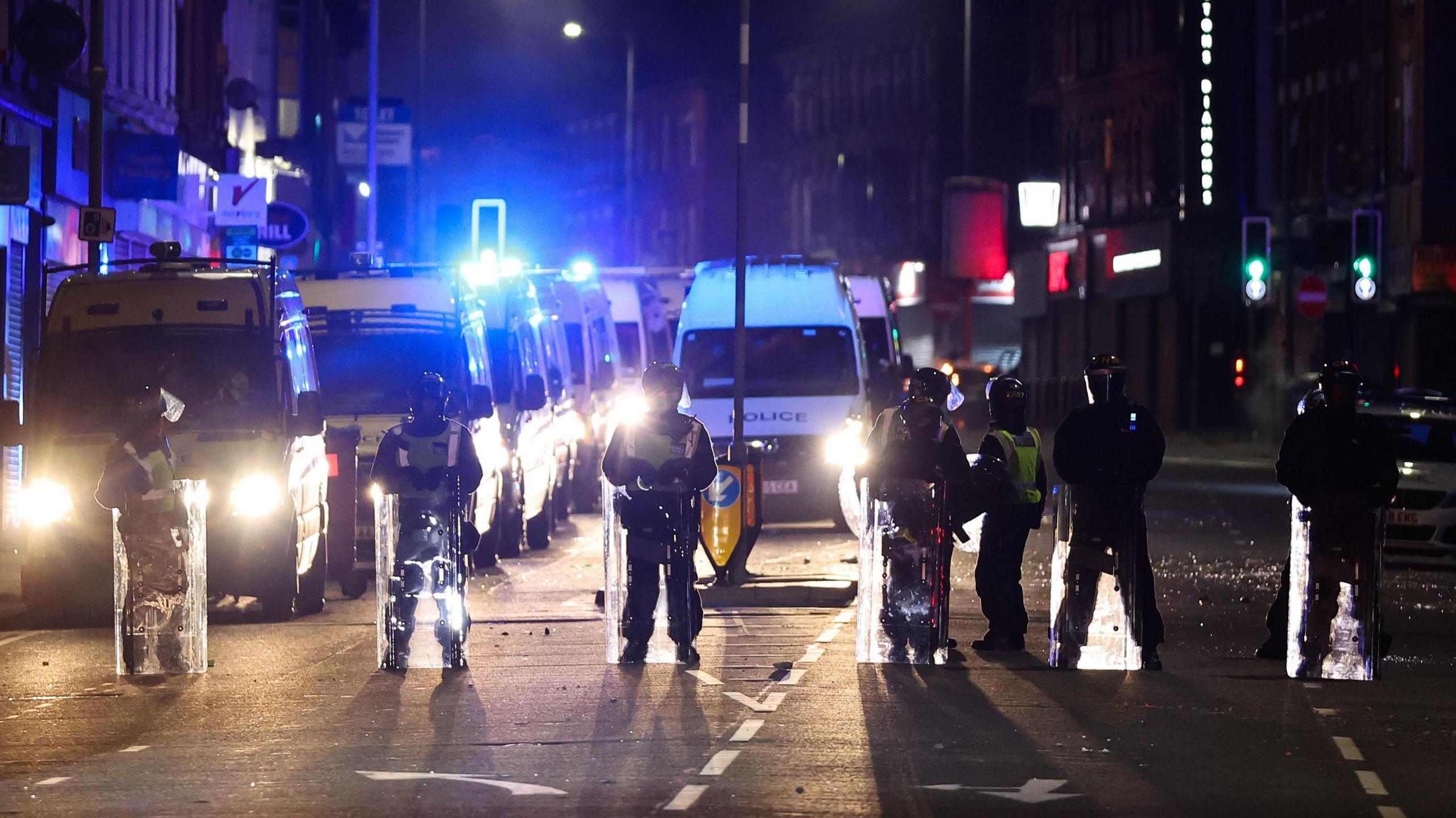 Riot police in helmets and shields line up outside closed shops during nighttime riots on County Road