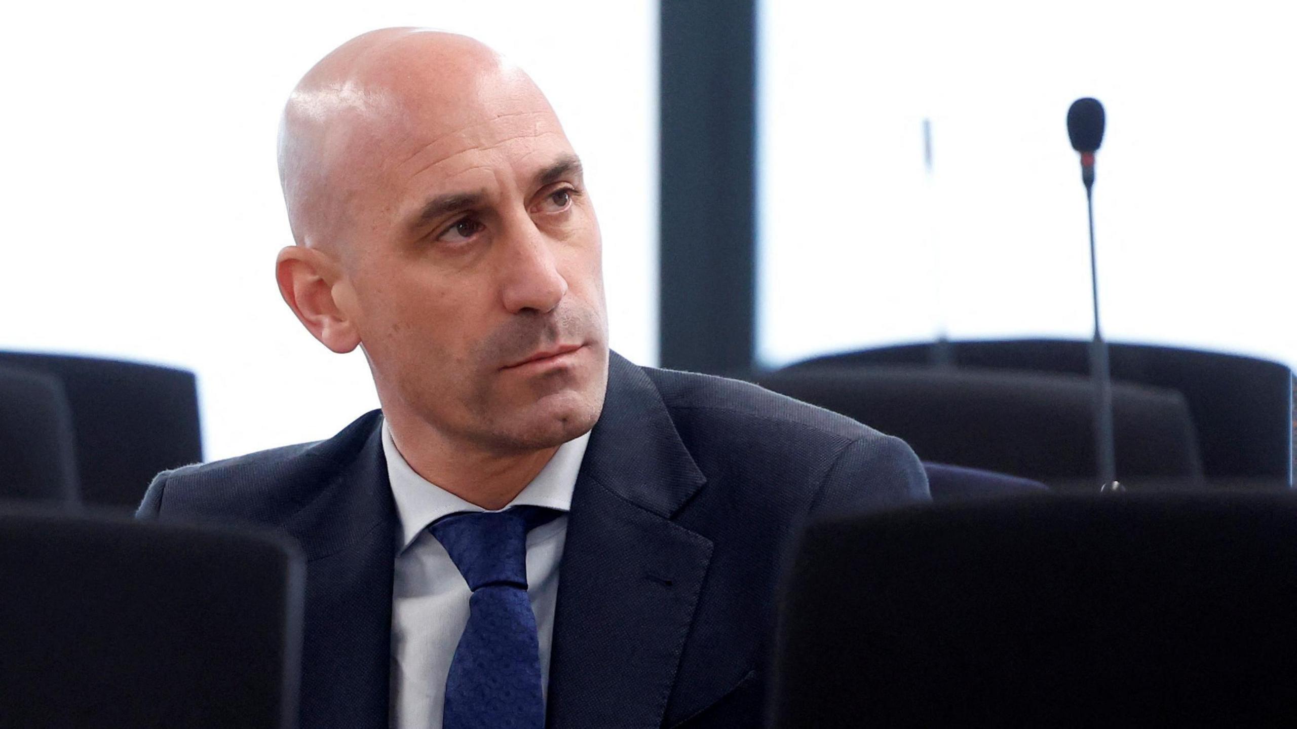 Luis Rubiales sits behind a desk in front of two large windows. He is bald and wears a dark blue jacket, white shirt, and blue tie.