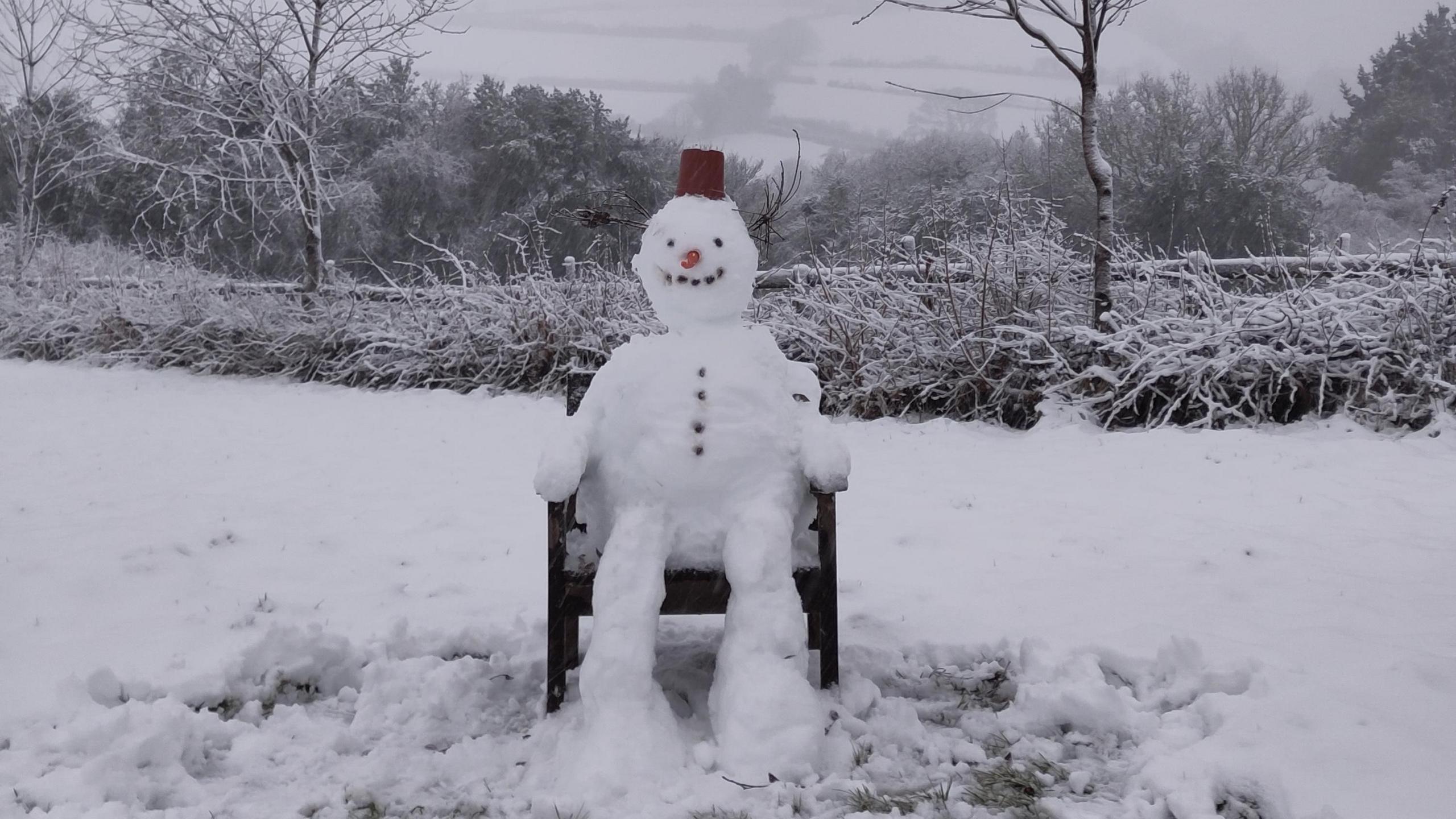 A snowman is sitting on a wooden chair in a  snow-covered garden. He is wearing a hat and has a carrot for a nose and stones for his eyes, mouth and buttons down his stomach.