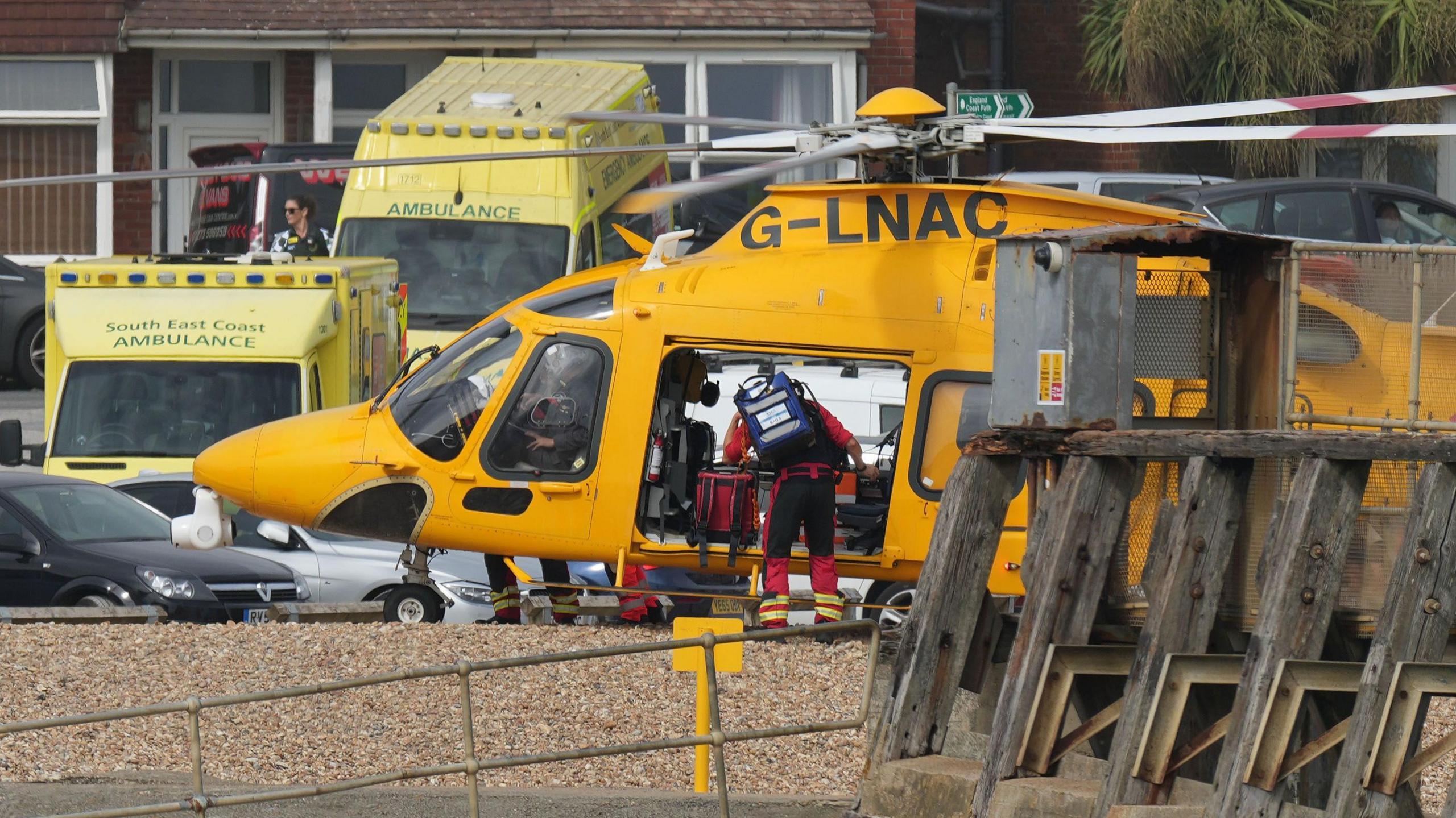An air ambulance helicopter is parked on the beach near Shoreham Port with ambulance vehicles and emergency personnel nearby.
