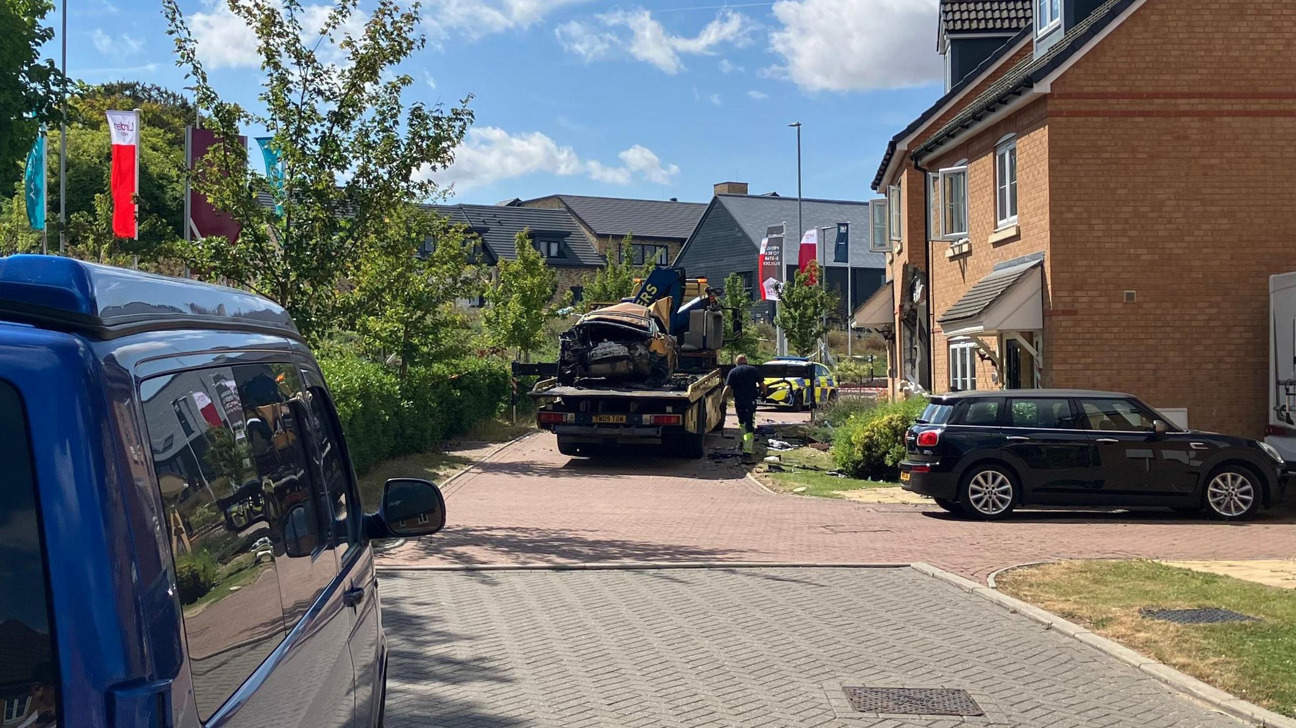 The destroyed car is on the back of a flatbed track close to the damaged house. It is a modern estate and it is a sunny day.