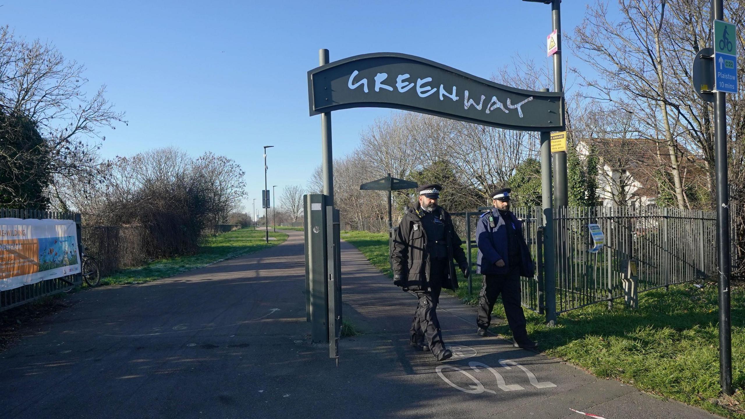 Police and Community Support Officers are pictured at the junction of the Greenway and High Street South in Newham, east London, where a newborn baby was found in a shopping bag by a dog walker. There is a large sign overhead with the words Greenway on it. There are trees and grass to the edge of the concrete path and a building in the background.