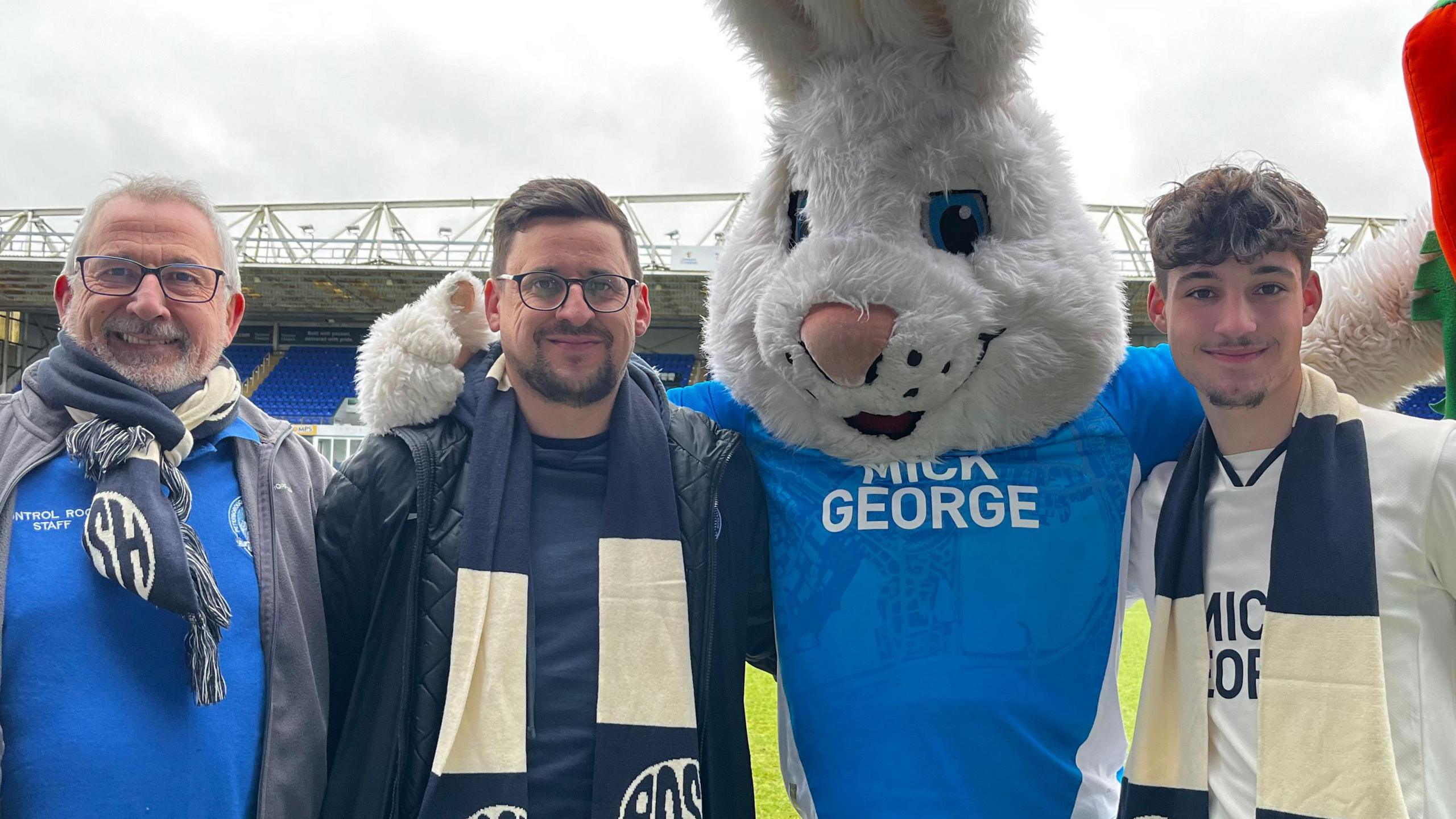 Michael Ferguson, his son, Ollie, Peterborough United mascot Peter Burrow and grandson Oscar, are stood in front of the pitch all with an arm around each other, wearing the club's branded T-shirts. Oscar, who is smiling at the camera, wears a white top with a blue and white scarf. He has brown hair, brown eyes and facial hair. Michael wears a blue top and scarf with black square framed glasses and a white shirt. He is smiling at the camera and has grey hair and grey facial hair. His son Ollie wears a black coat, blue top, blue and white scarf and circle framed glasses. He is smiling and has brown hair and facial hair. 