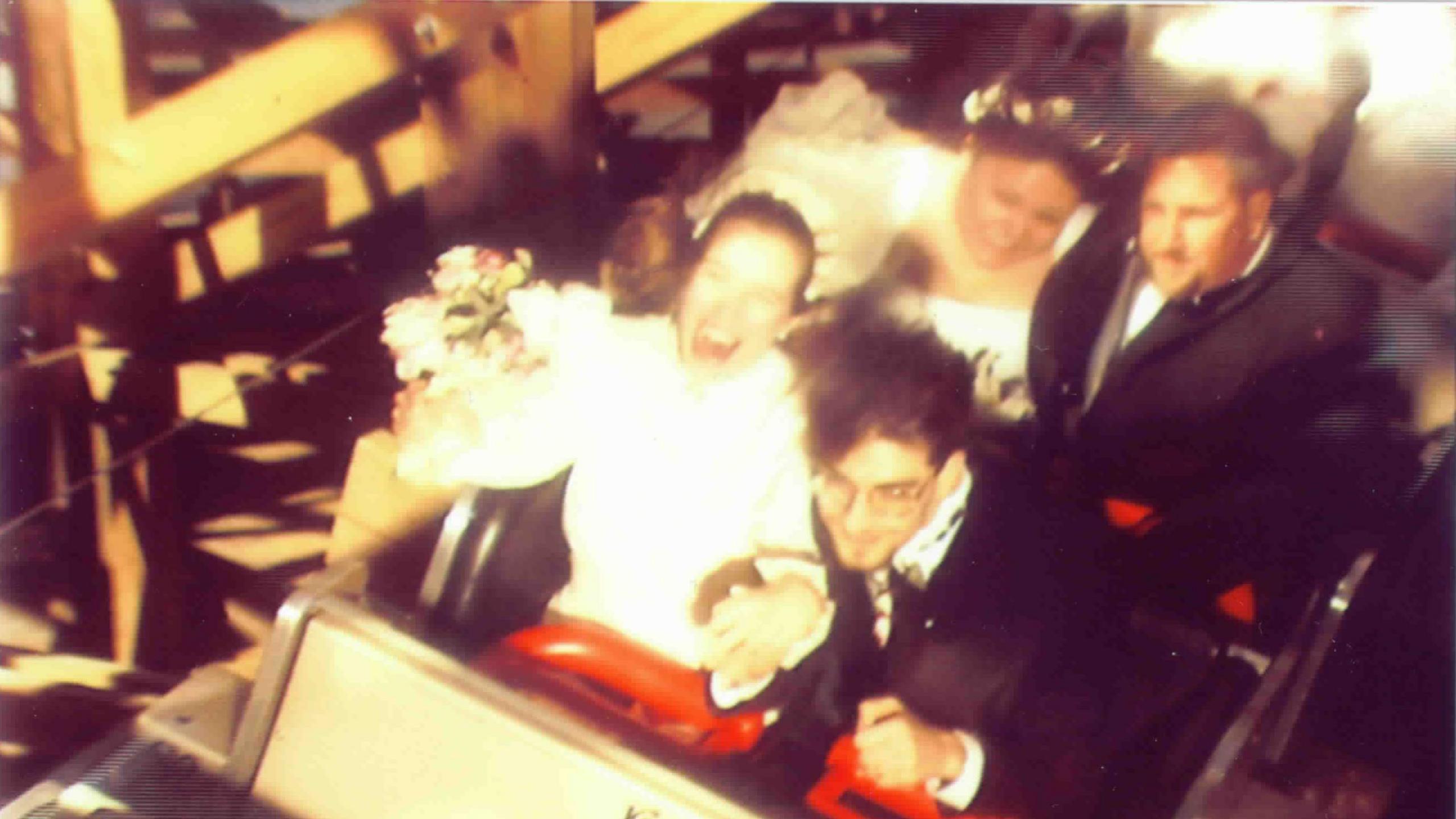 Korey and his wife in the front cart of a rollercoaster. He is wearing a black suit, she is wearing a white wedding dress and holding a bouquet. In the row behind is another man in a dark suit and a woman in a light dress with a flower headband. 