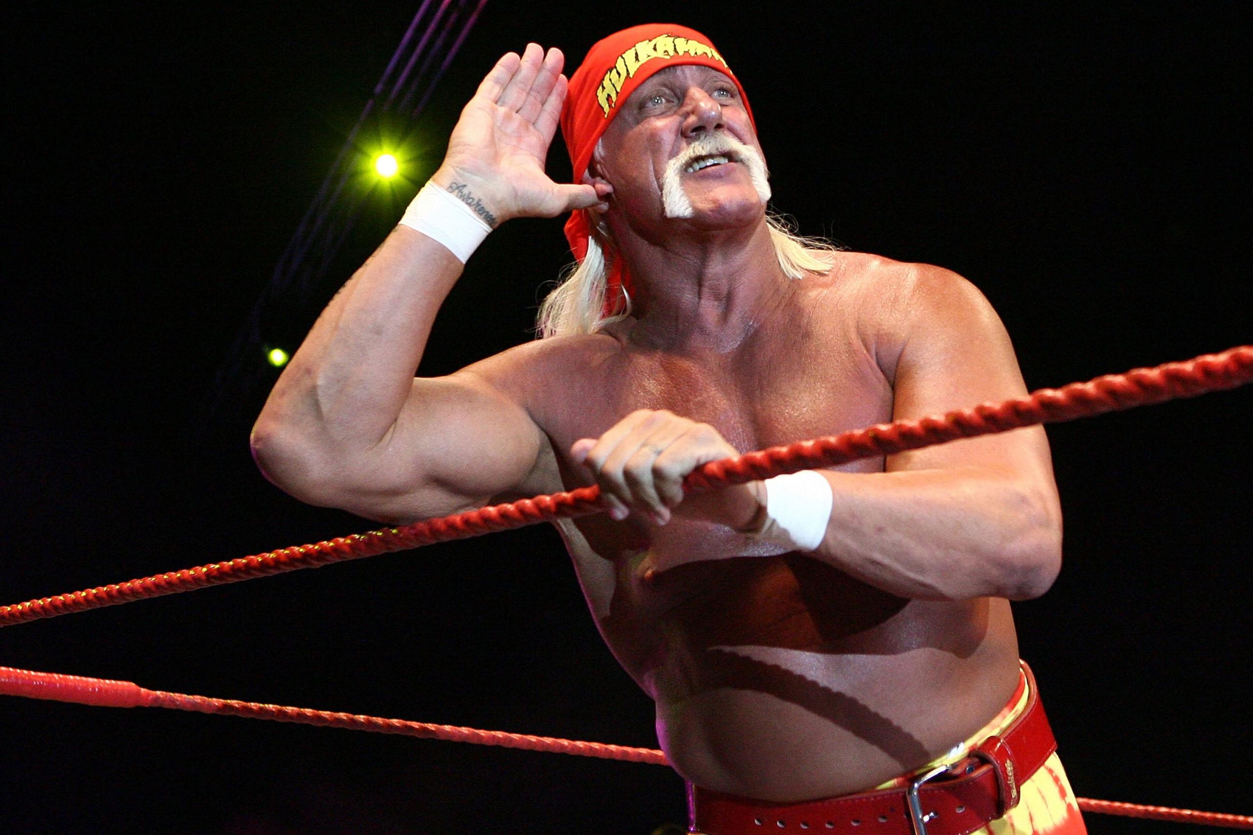 Hulk Hogan është brenda një arene mundjeje, duke u mbështetur në litarët e kuq me njërin krah, ndërsa ngre dorën tjetër te veshi në një gjest dëgjimi. Ai mban veshur një shami të kuqe të ndezur, mbathje të kuqe të kombinuara me një rrip të verdhë dhe ka flokë të gjatë e të çelët që i zbresin poshtë shpinës. Sfondi është i errët me reflektorë që ndriçojnë skenën, duke krijuar një atmosferë dramatike tipike për një garë profesionale mundjeje.