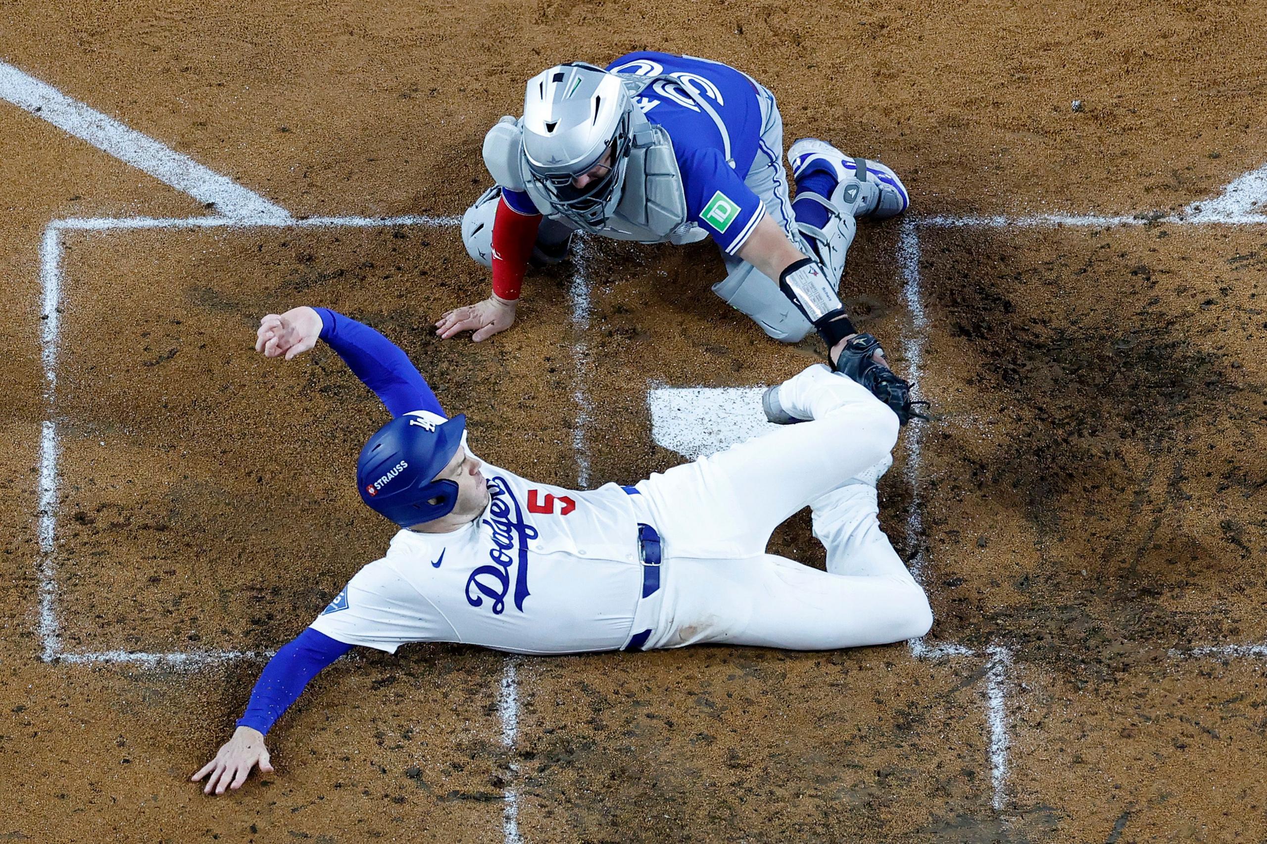 A baseball player in a white Dodgers uniform slides across home plate on a dirt field, extending one arm toward the base. A catcher in gray and blue gear crouches nearby, reaching forward as the ball remains out of view.