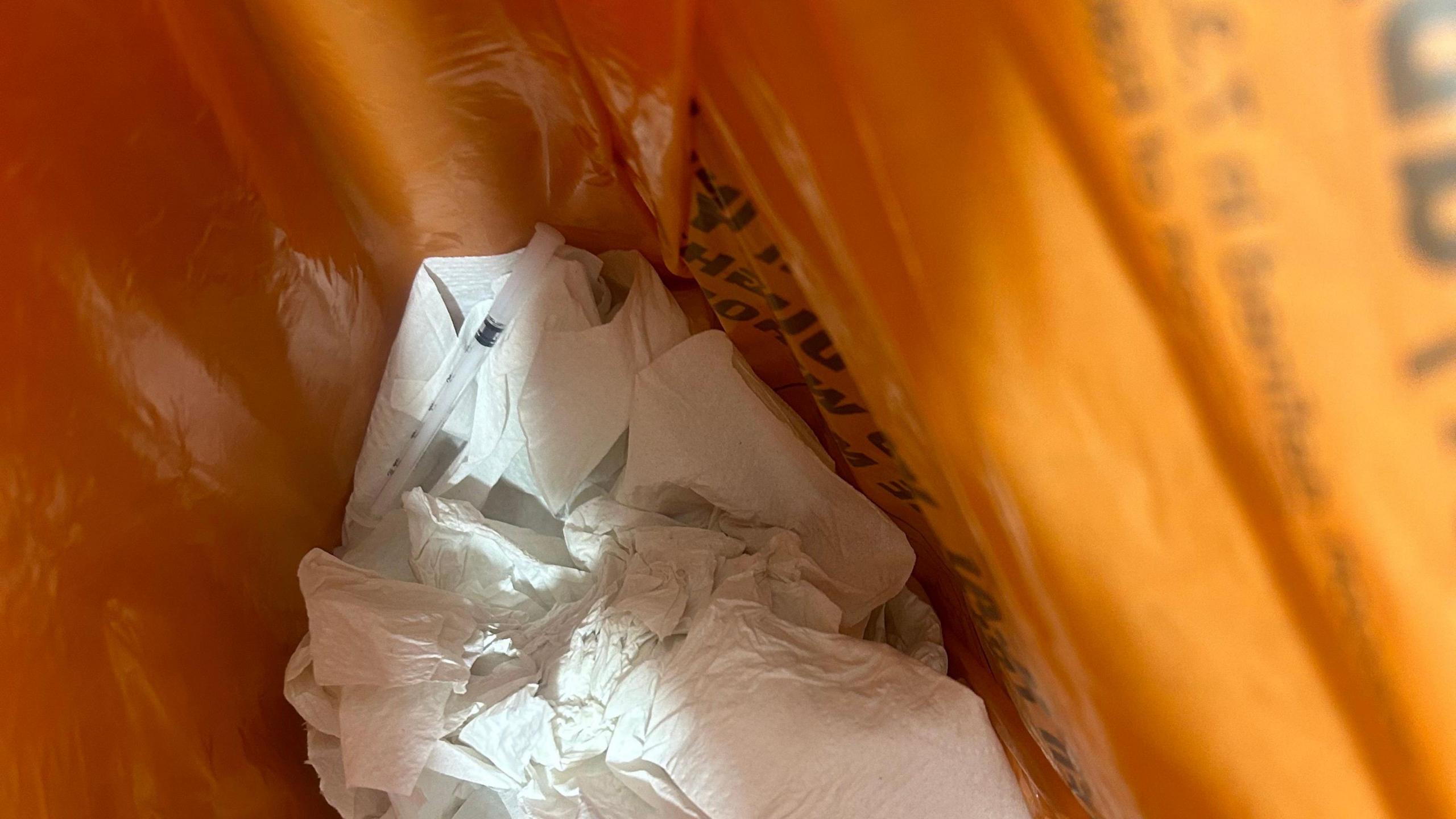 Small thin needle beside a wad of paper in an orange bin bag