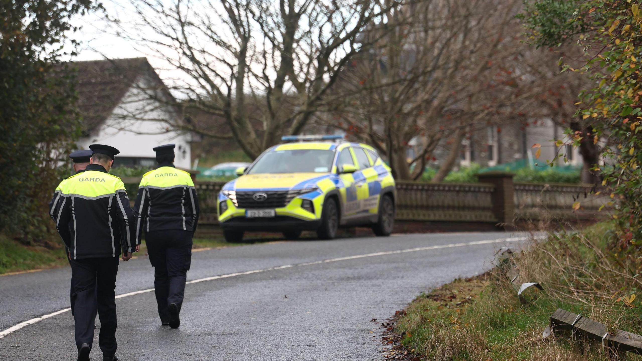 A Garda patrol car is parked at a road junction with two Garda officers standing nearby.