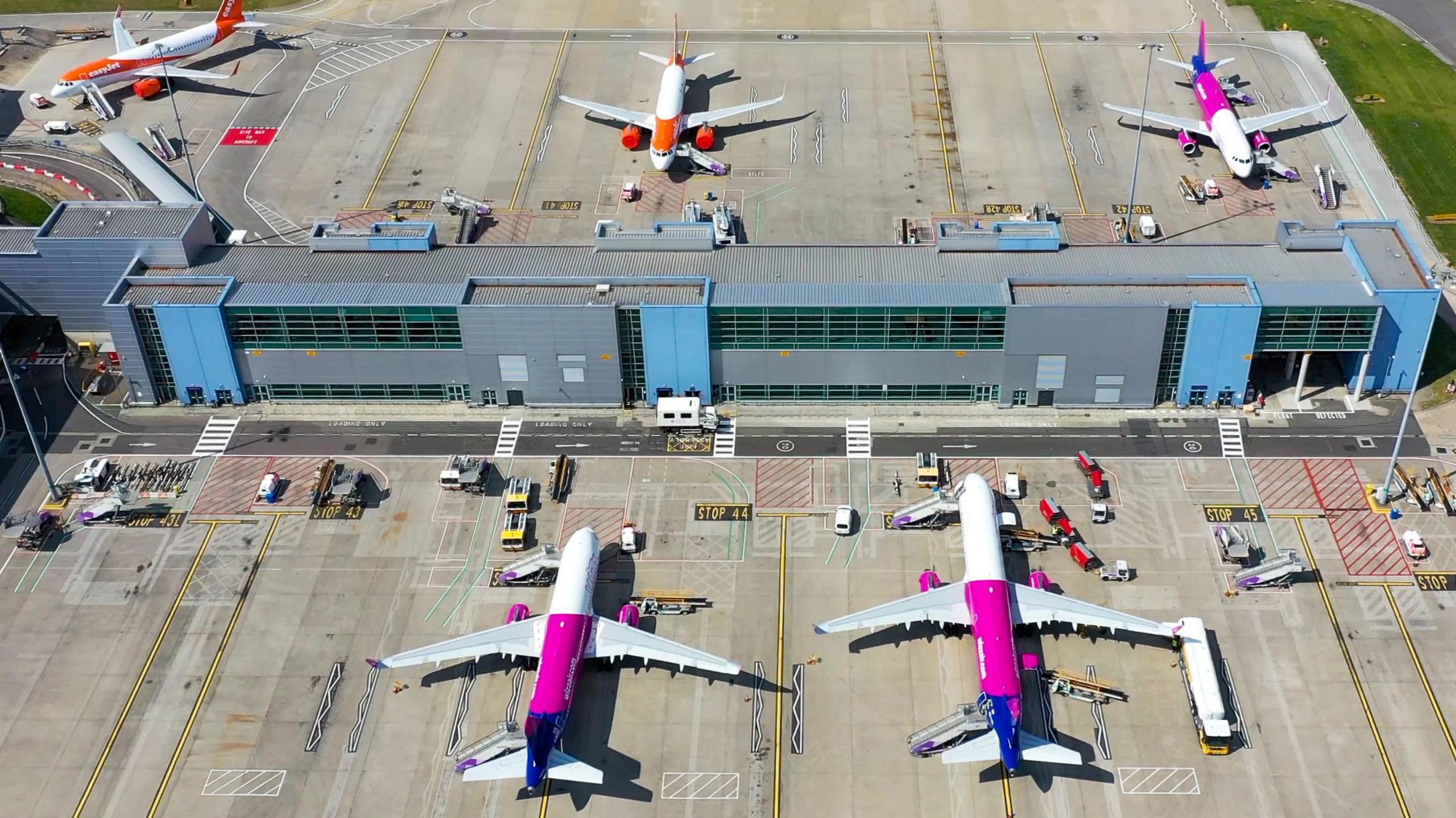 An aerial view of Luton airport, showing five planes on the ground, a large terminal building, other vehicles are on the ground. There is grass to the right, and a large concrete ground.