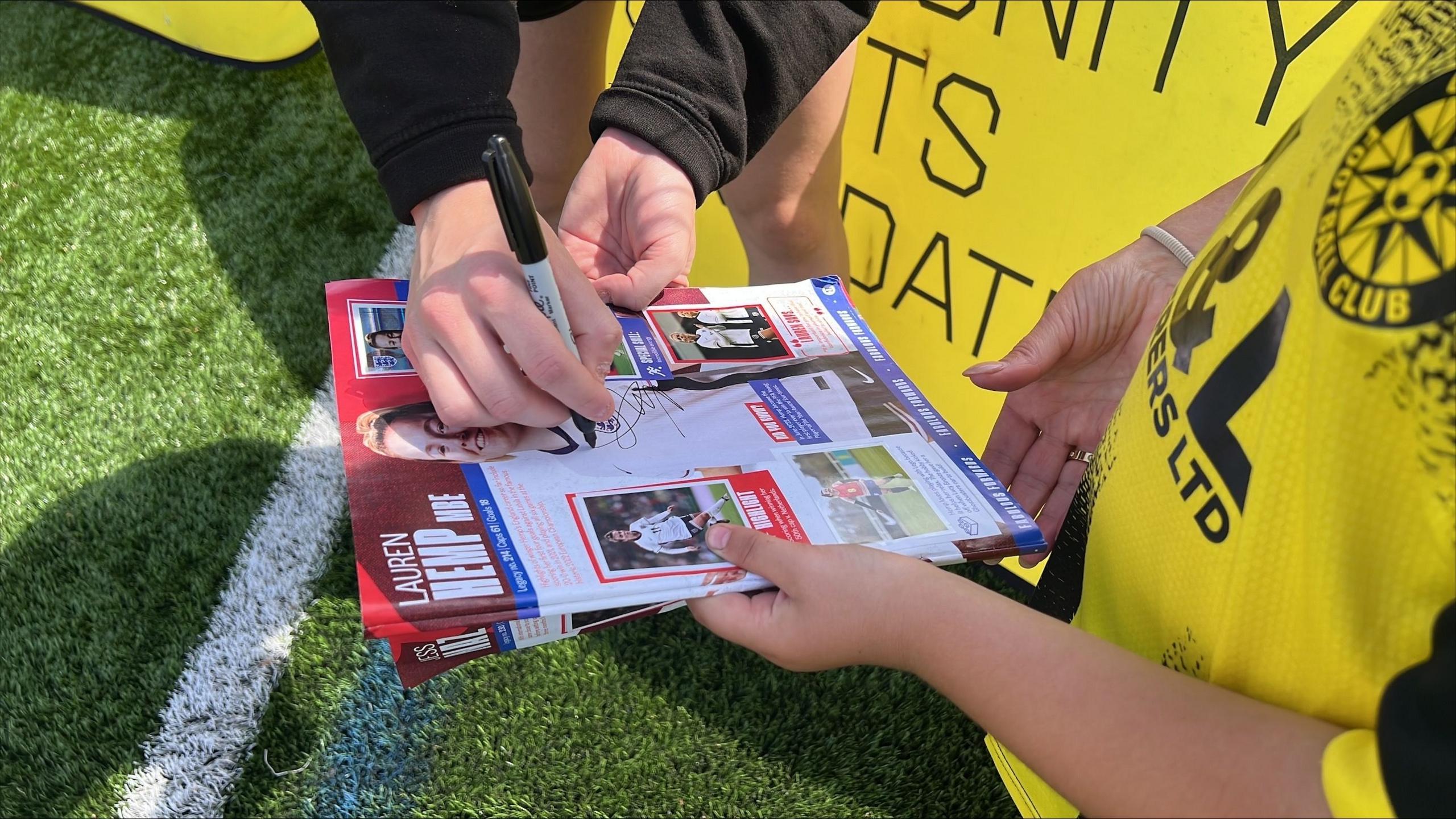 Lauren Hemp's hand can be seen, as she signs a leaflet or programme, which features a picture of herself on the front. A younger girl is holding the leaflet.