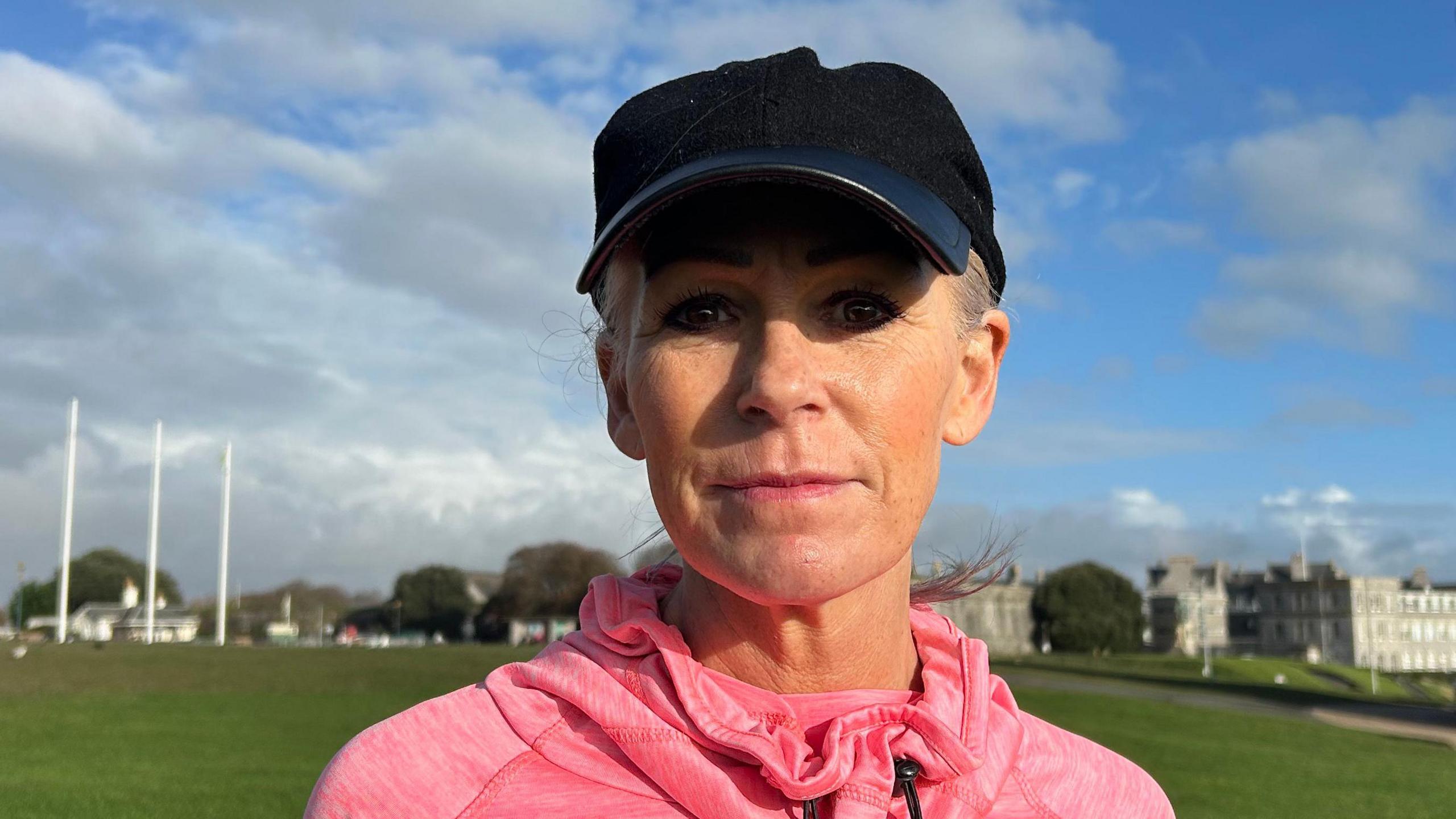 Nicky Putman is wearing a pink running jumper and a black cap on a bright sunny day in Plymouth. She is stood on grass and looking at the camera. There is buildings and flag posts in the background. 