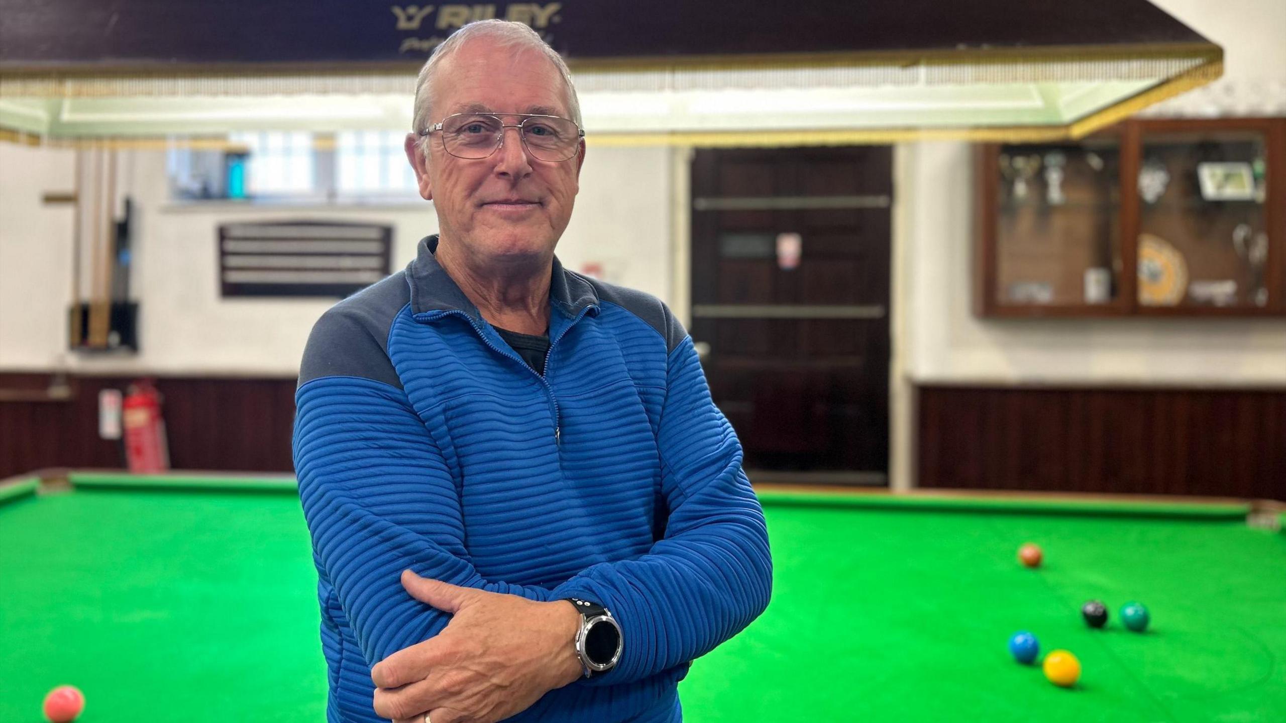 A man looks directly towards the camera standing side on. He is stood in front of a large green snooker table with multicoloured snooker balls on top. He wears a blue pullover jumper with dark patches on the shoulder and neck. A large black smart watch is on his right arm.
