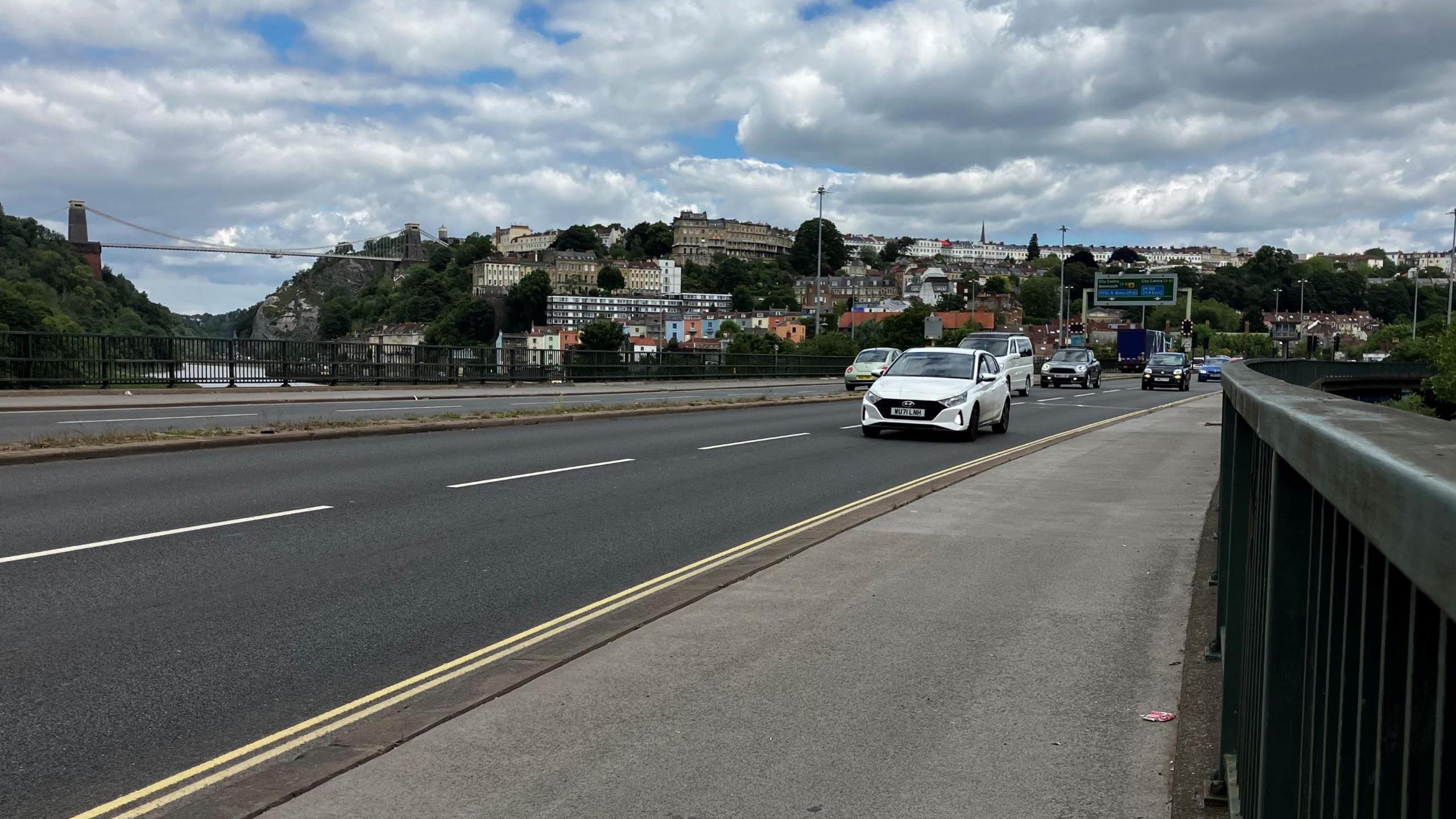 Cars are seen driving on Brunel Way in Bristol, with the Clifton Suspension Bridge and houses in Clifton visible in the background