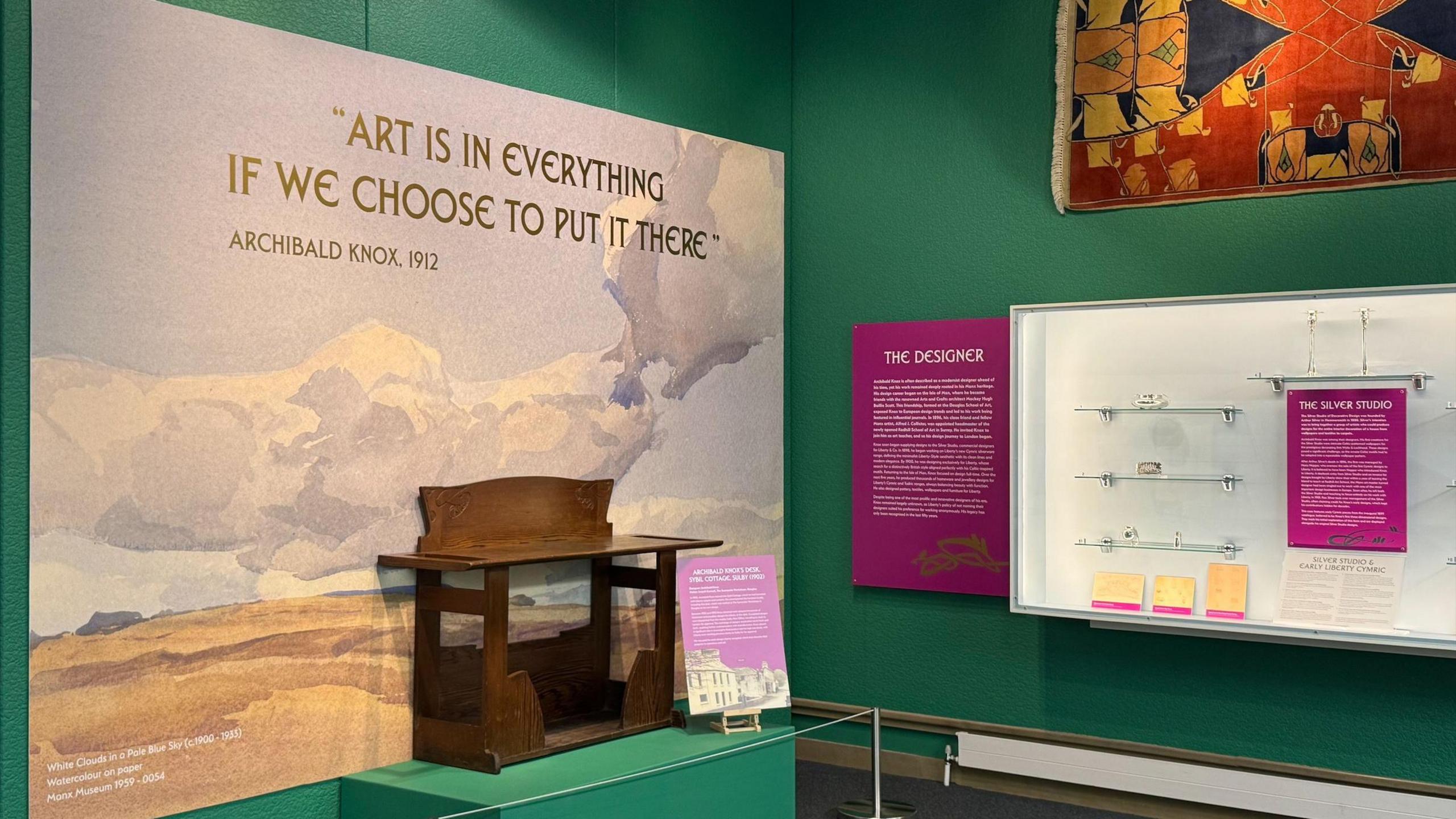 An area of the exhibition, with a large watercolour landscape, a brown wooden bench and a glass cabinet with silverware. There is also a brightly coloured rug attached to the wall.