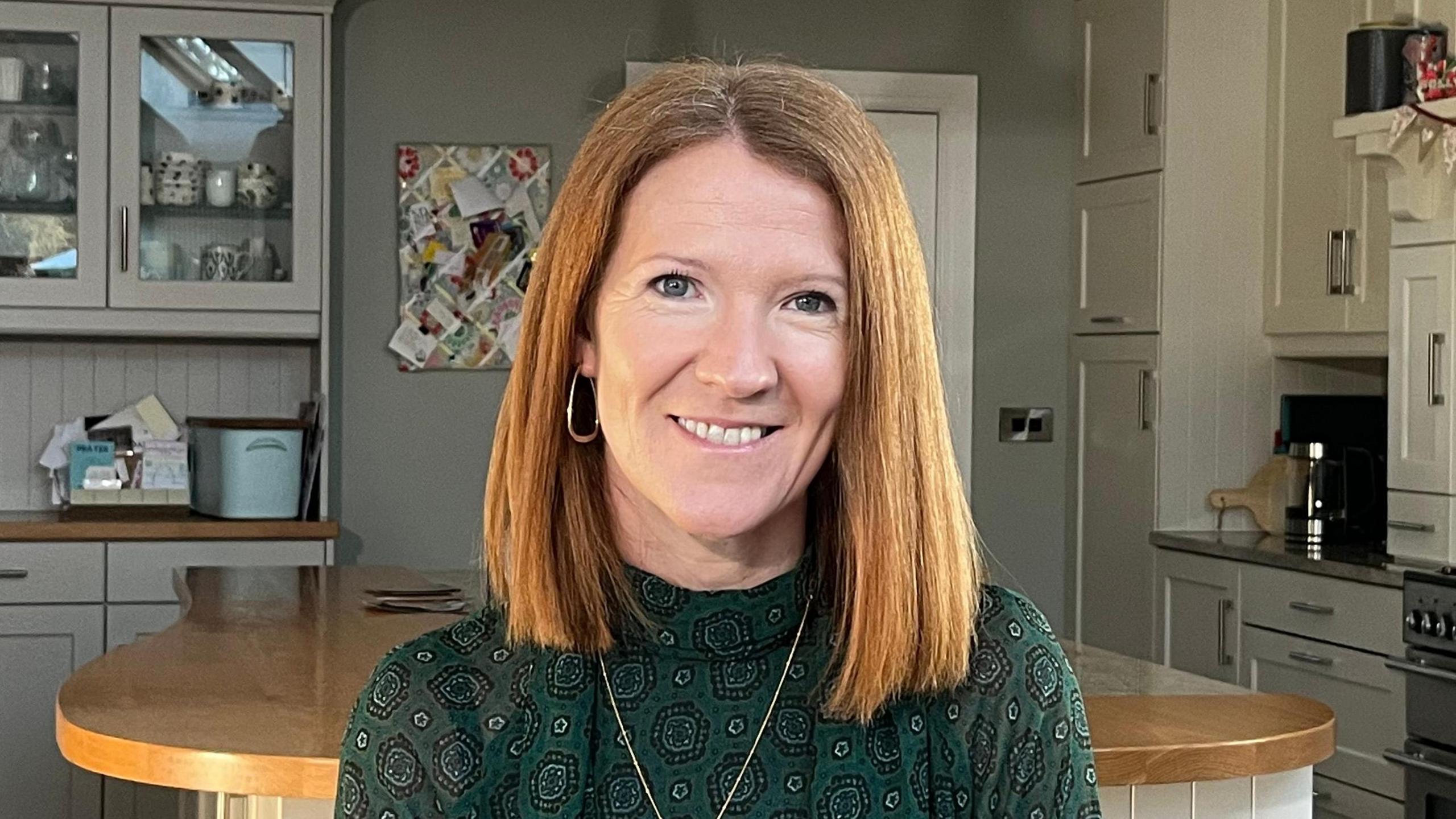 Angela smiling. She has shoulder length light auburn hair. She is wearing a green patterned top and a gold necklace. She is also wearing gold hoop earrings.