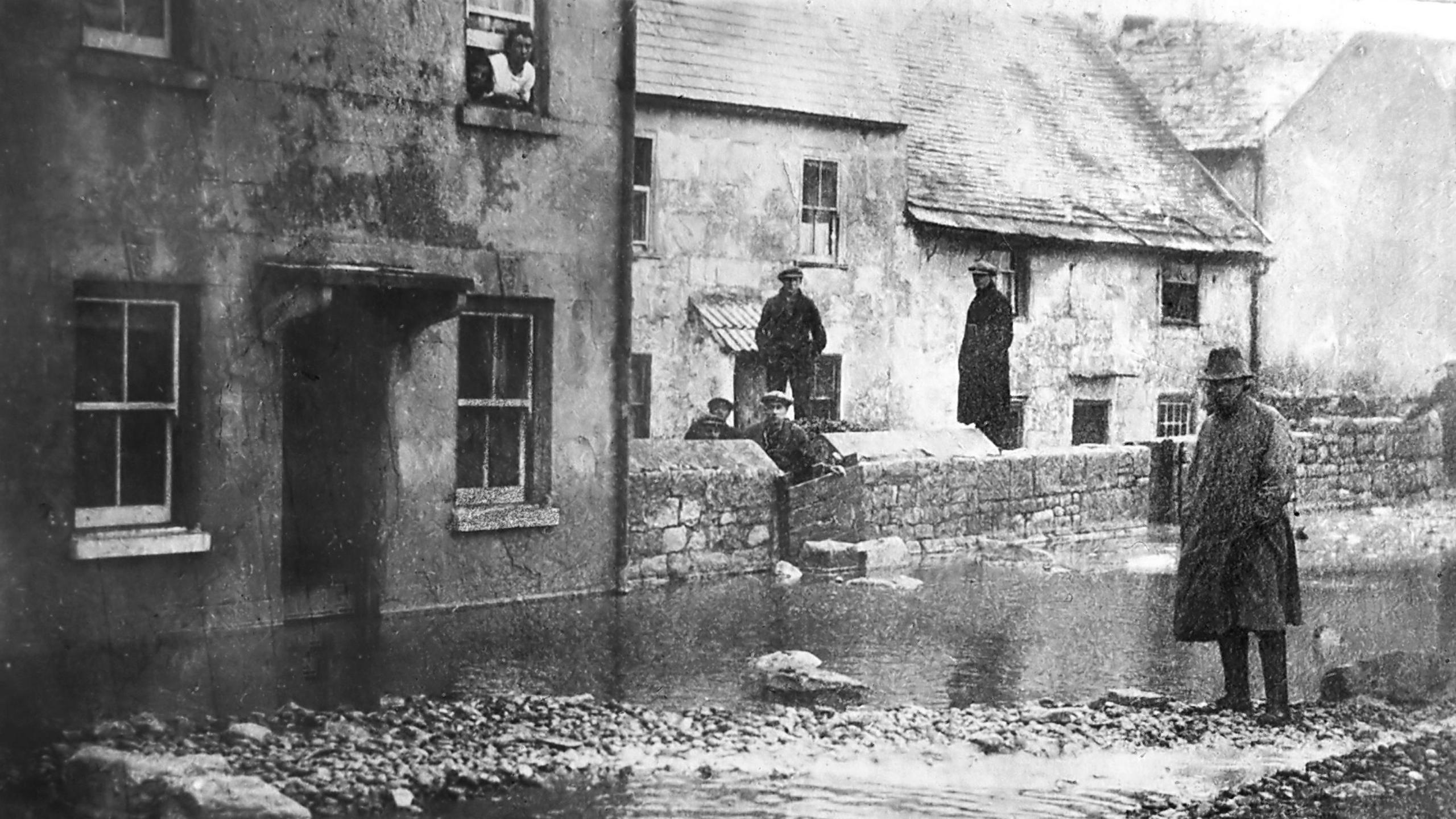 Black and white photo from November 1924 shows tidal wave flooding in Chiswell. A man wearing a raincoat and a trilby hat is standing in a flooded street lined with terraced cottages. Five men are standing behind the front wall of one of the houses and another is leaning out of a first-floor sash window.