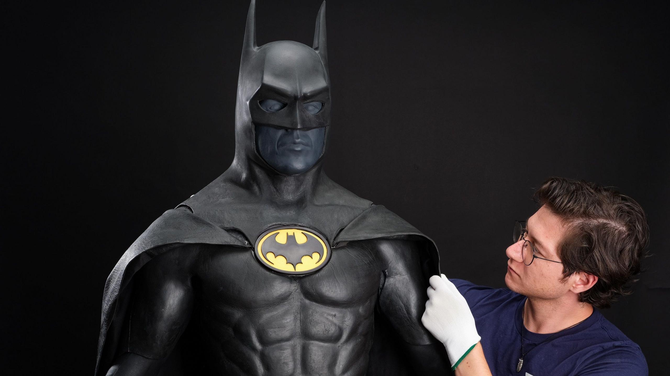 A man wearing glasses and white gloves adjusts the cape on a black Batman suit, which has the Batman Logo in a yellow circle displayed on its chest.