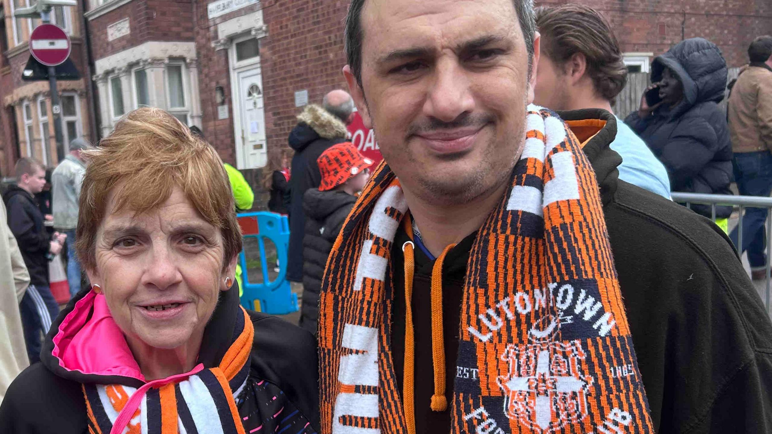 Rosie Moore has reddish short hair, and is wearing earrings, a black coat with a hot p9ink lined hood and an orange, white and black Luton Town scarf and her son has black hair, a moustache and is wearing a black hoodie and Luton Town scarf too.