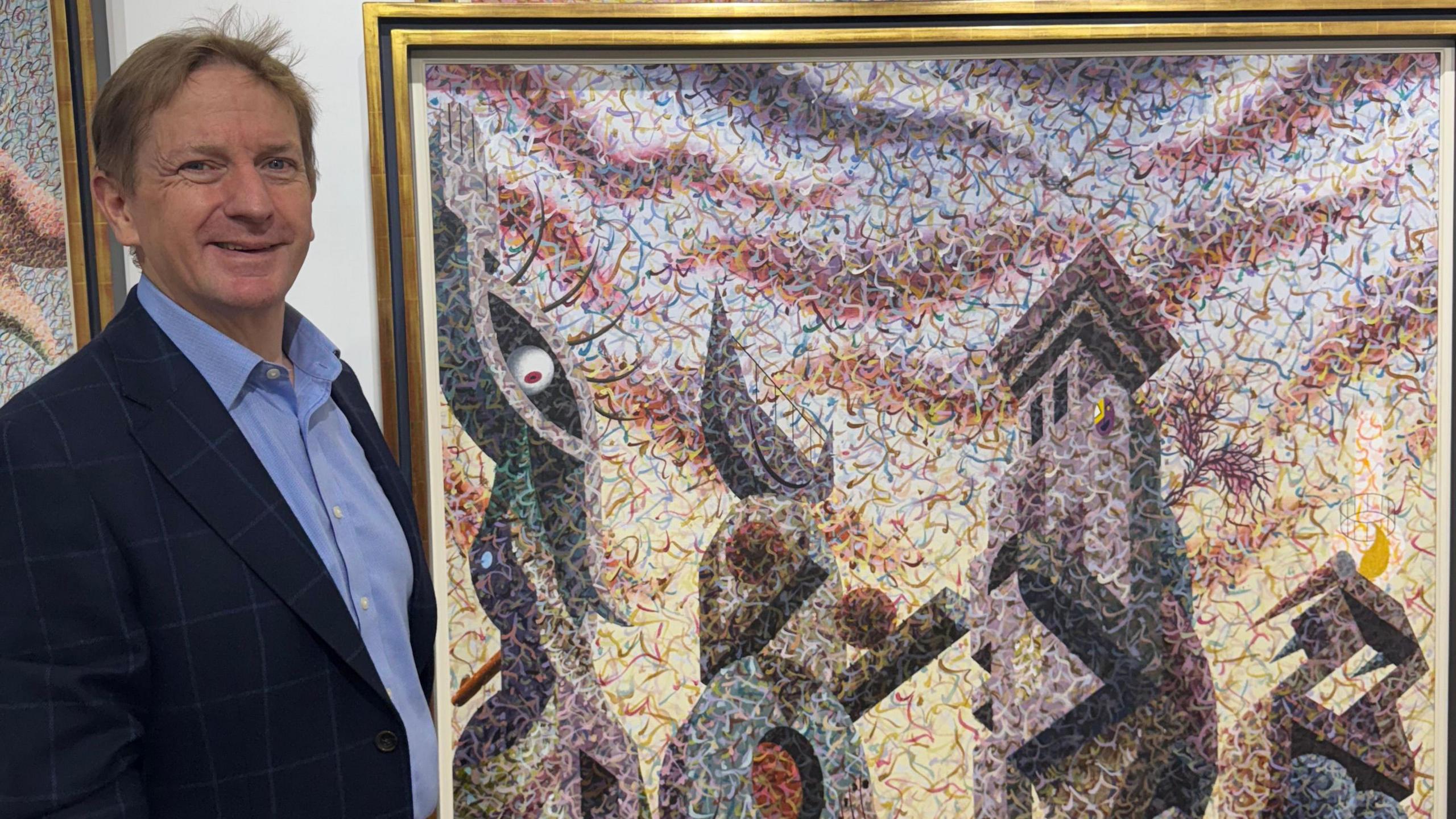 Grant Ford smiles looking into the camera while standing next to a large surrealist painting, featuring towers and an eye