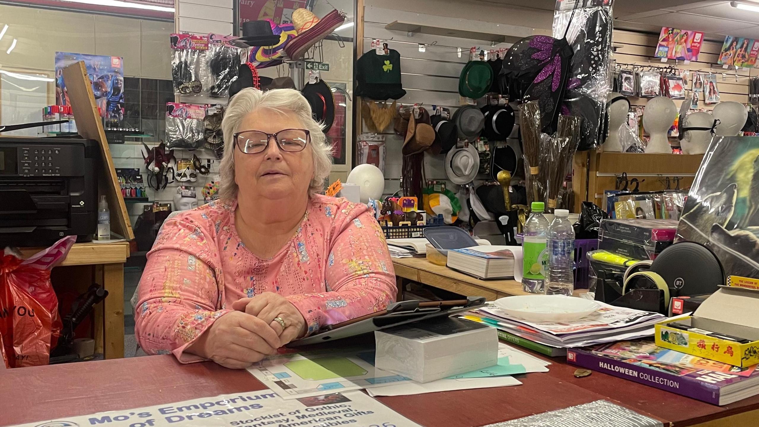 A woman sits behind a counter in a novelty item shop. She is wearing a pink top and has glasses. Behind her are a number of hats and dressing-up items. 