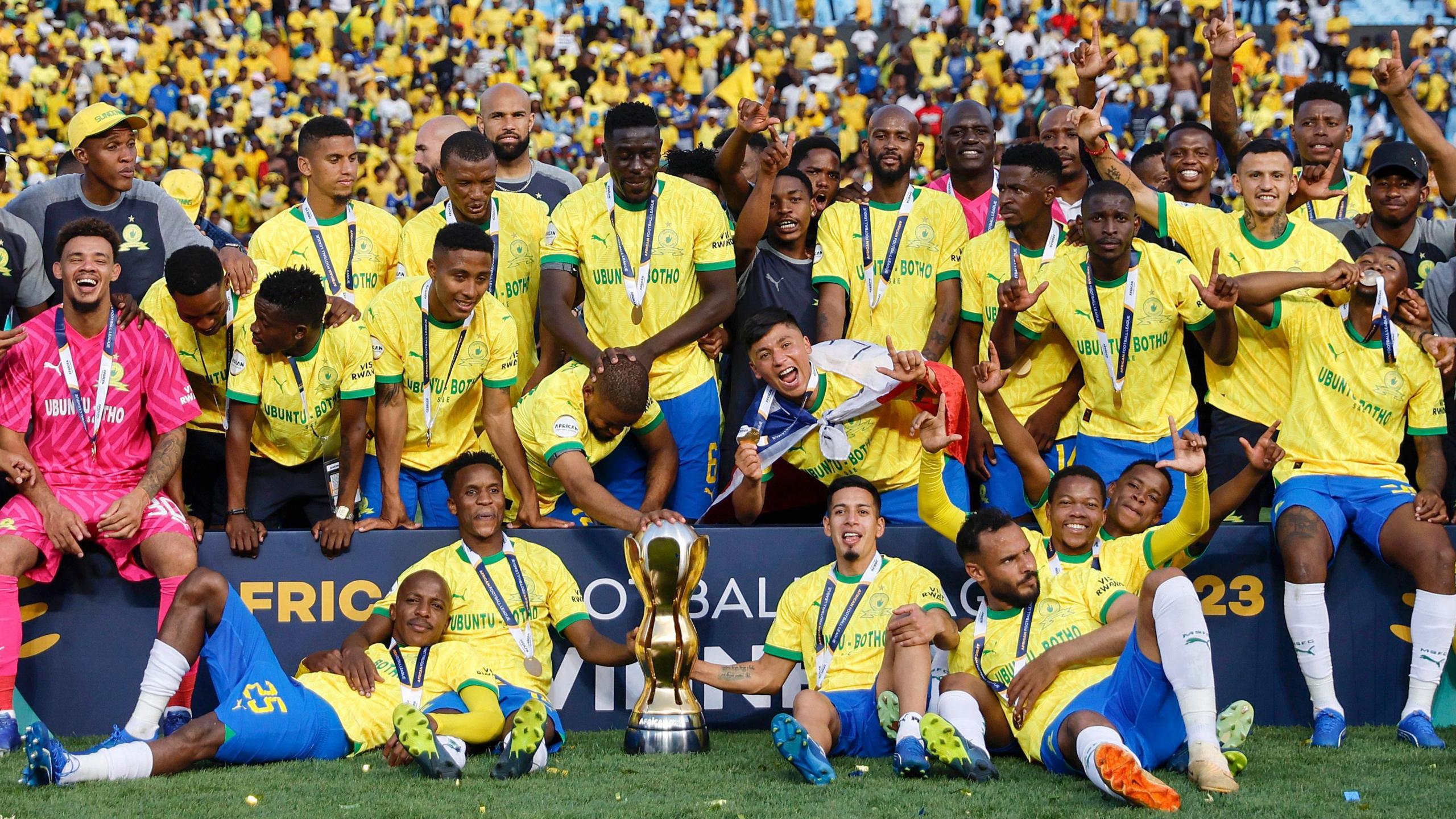 A group of Mamelodi Sundowns players, predominantly wearing yellow shirts, blue shorts and medals around their necks, crowd around an advertising hoarding as they celebrate with the African Football League trophy