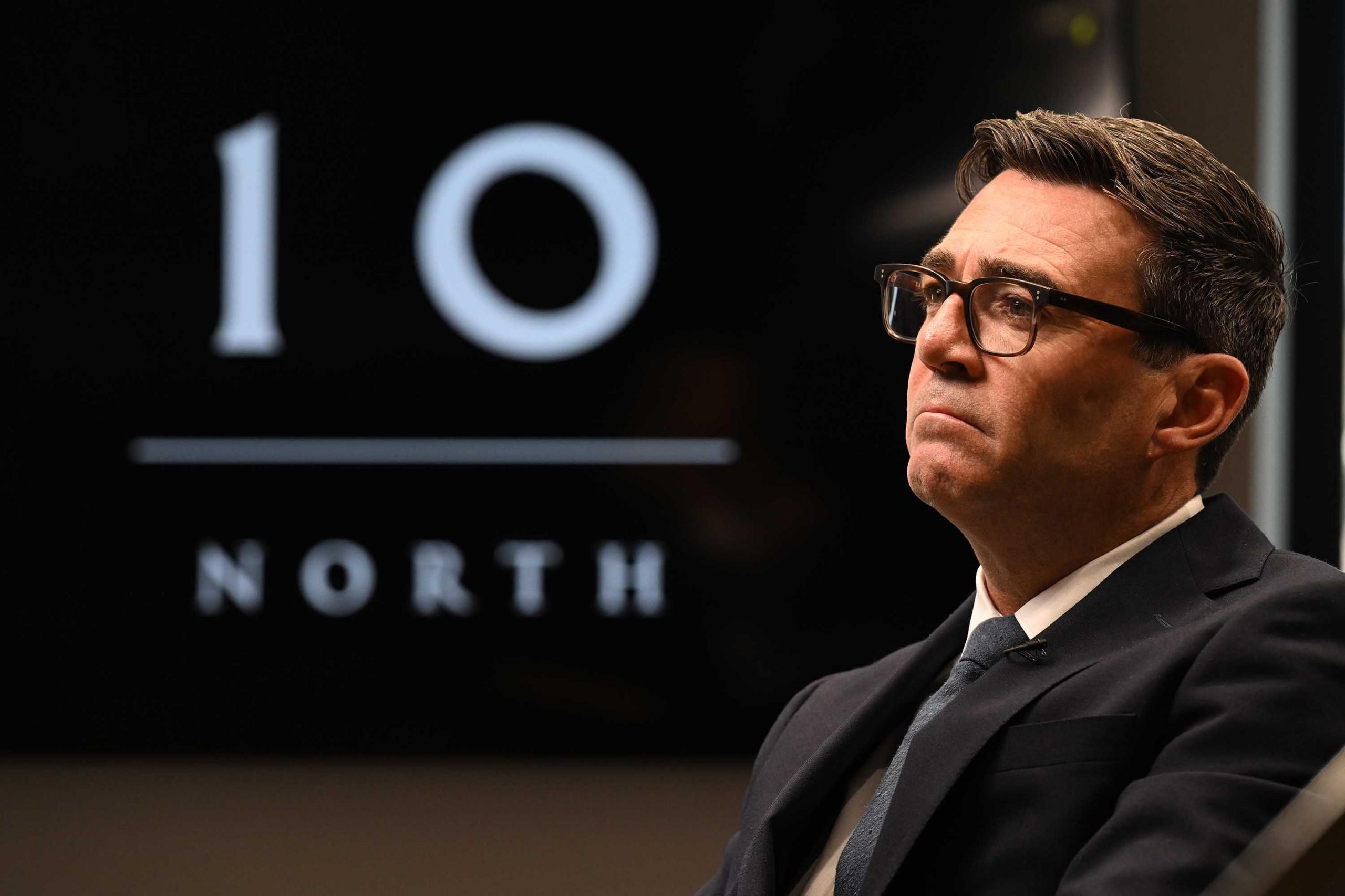A man in a dark grey suit and dark blue tie white shirt with black glasses in front of a number 10 and north