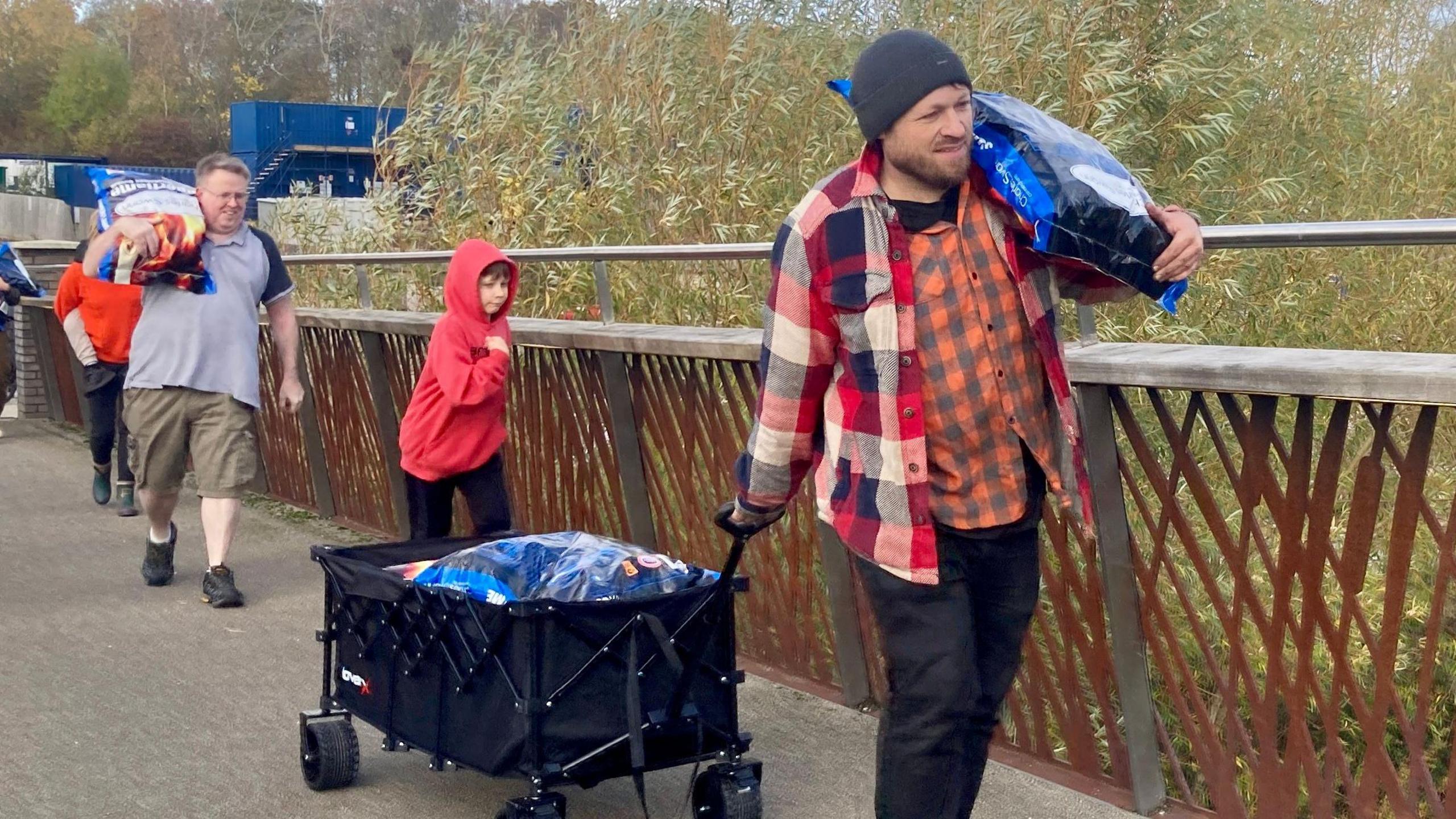 Andy Simpson, wearing an orange checked shirt under a red and blue overshirt. is dragging a cart full of coal bags behind him, with another blue bag balanced on his shoulder.